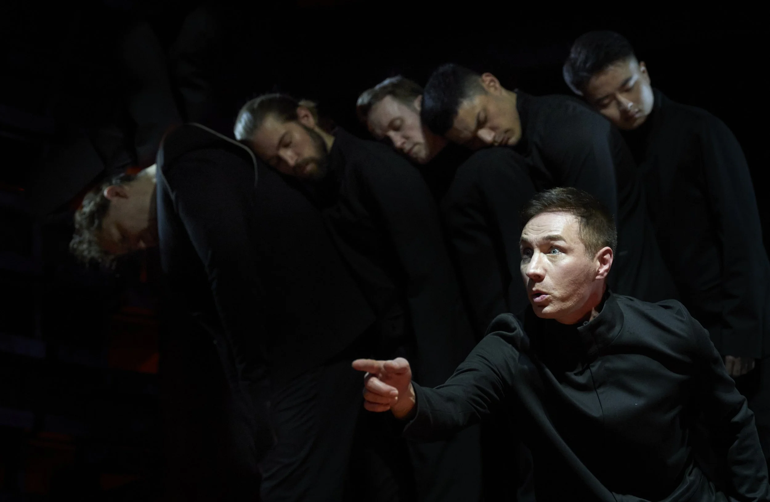 Ein Mann im schwarzen Hemd steht im Vordergrund und spricht dramatisch, während im Hintergrund fünf Personen in dunklen Kleidern mit geschlossenen Augen stehen und den Kopf nach unten geneigt haben.