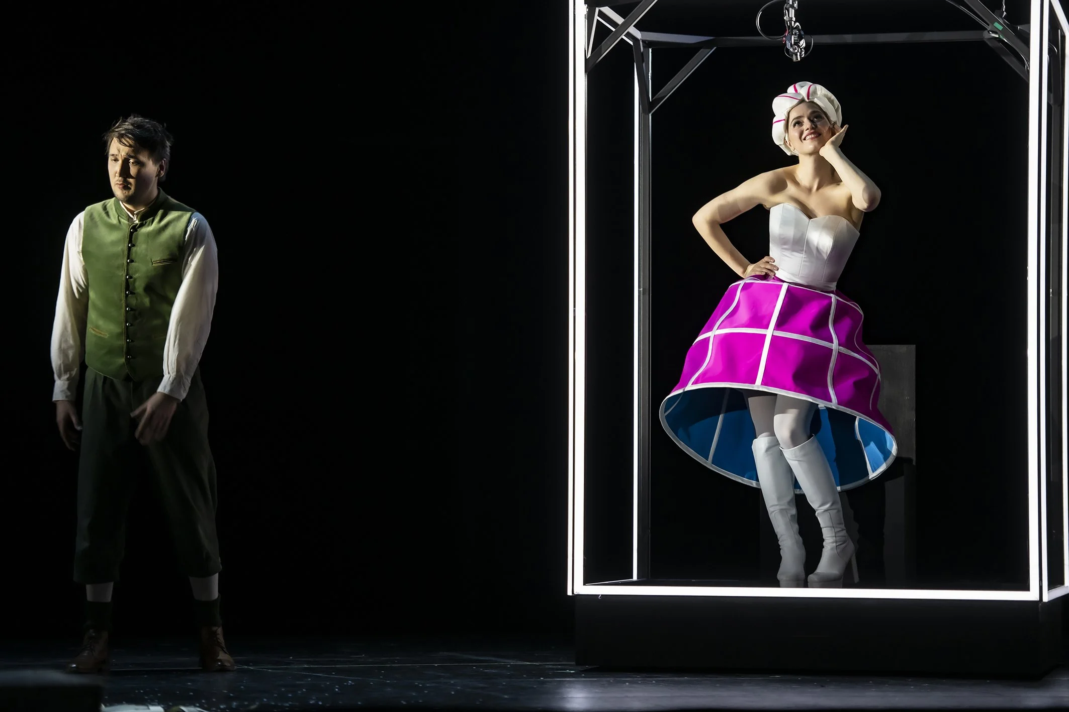 Zwei Schauspieler auf einer dunklen Bühne, einer steht links in traditioneller Kleidung, die andere rechts in einem bunten Bühnenkostüm in einer Glasvitrine.