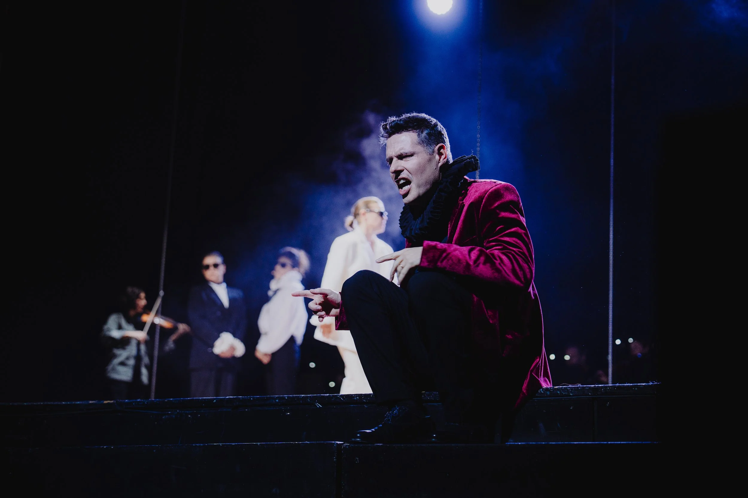 Schauspieler in dramatischer Szene mit mehreren Darstellern im Hintergrund, unter starker Beleuchtung, auf einer Theatbühne während einer Aufführung.