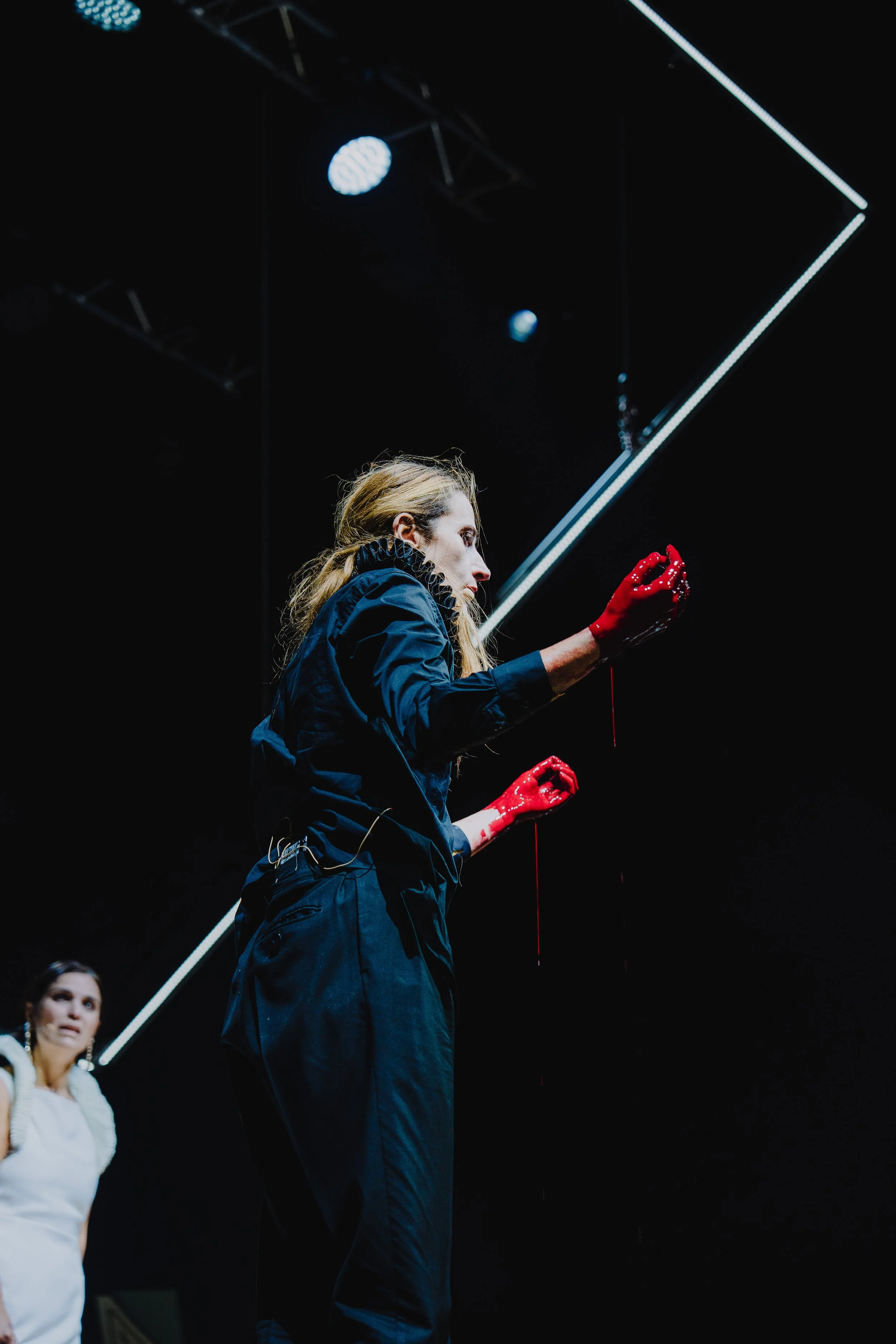 Eine junge Frau steht auf einer Bühne, trägt schwarze Kleidung, rote Handschuhe und schaut nach oben. Im Hintergrund ist eine zweite Person zu sehen, die ebenfalls auf der Bühne steht.