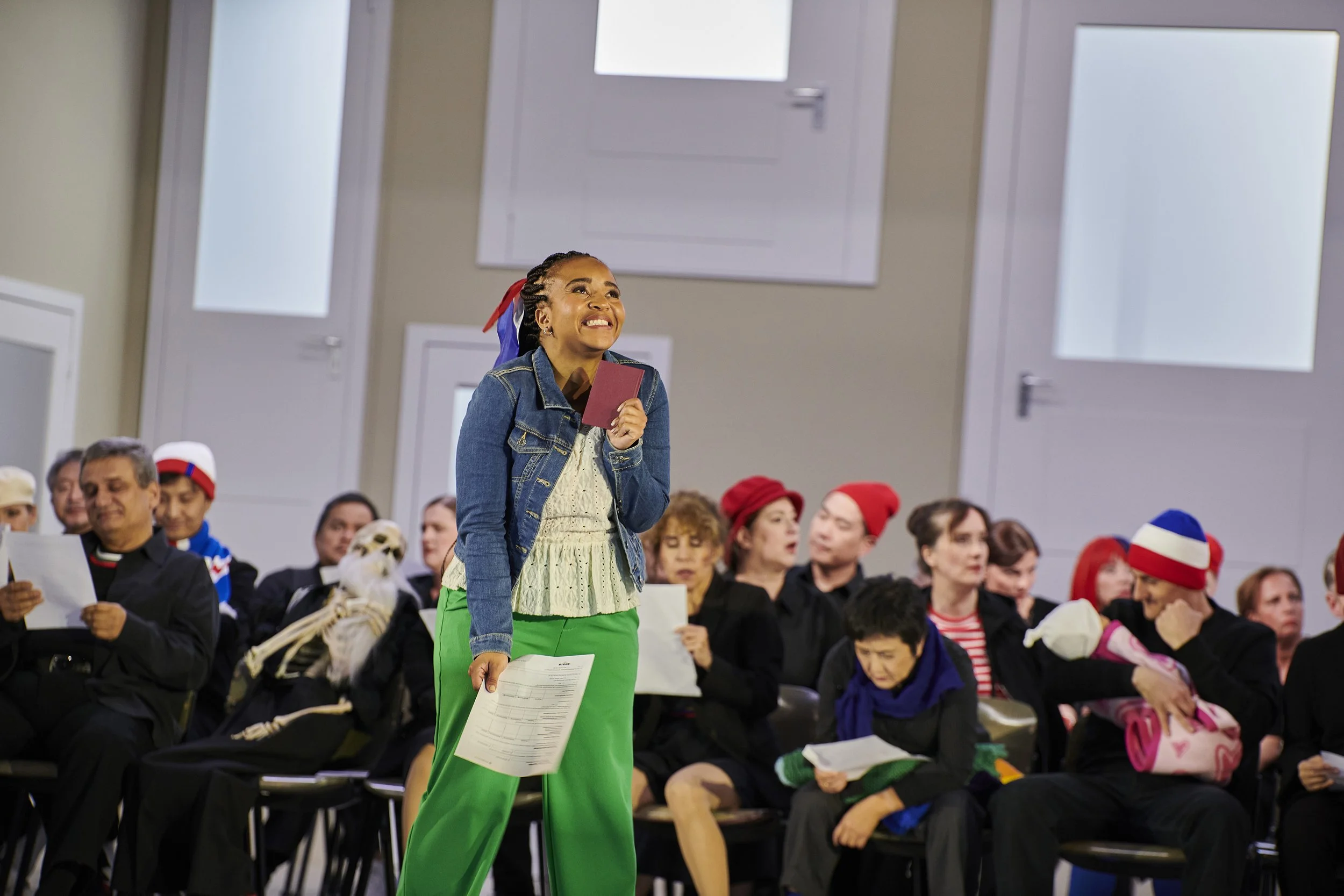 Eine junge Frau in bunter Kleidung steht lachend in einem Wartezimmer, umgeben von anderen wartenden Menschen. Einige tragen Hüte und Halstücher, einige lesen Papiere.