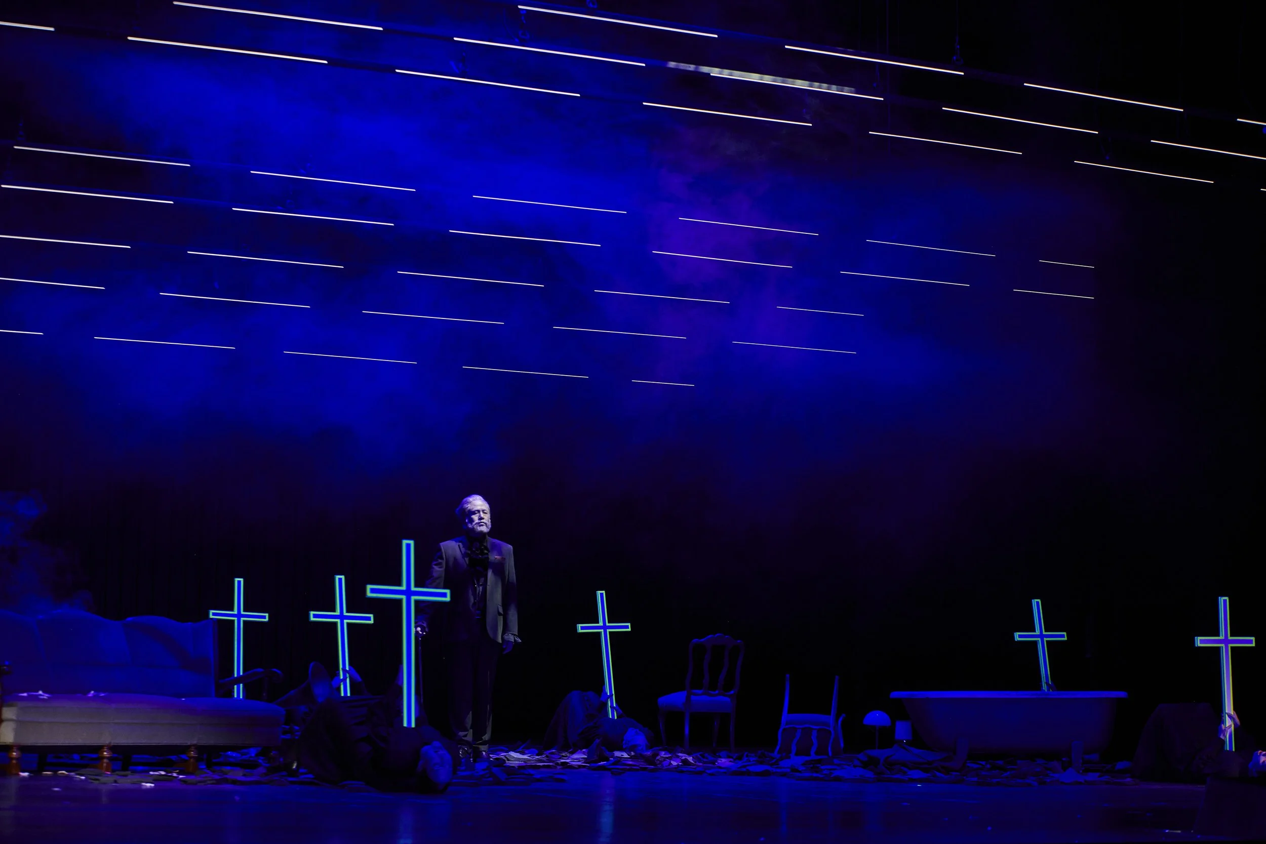 Eine Theaterszene mit blauer Beleuchtung, mehreren leuchtenden Kreuzen, einem Sessel, einem Bett und einer Person in dunkler Kleidung auf dem Boden, während eine andere Person steht.