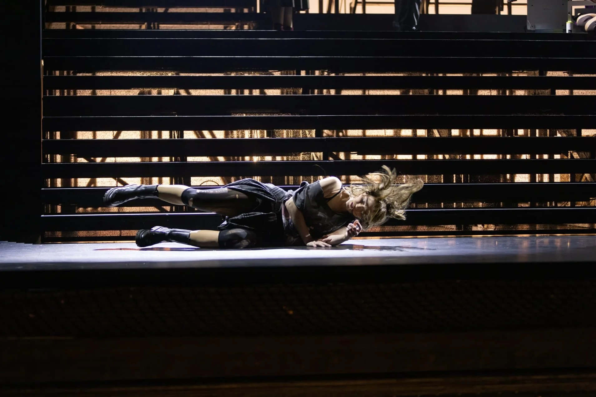 Eine Person liegt auf dem Boden vor einer dunklen, schrägen Holzstruktur, beim Tanzen oder Performing auf einer Bühne, mit verrücktem Haar und schwarzer Kleidung.