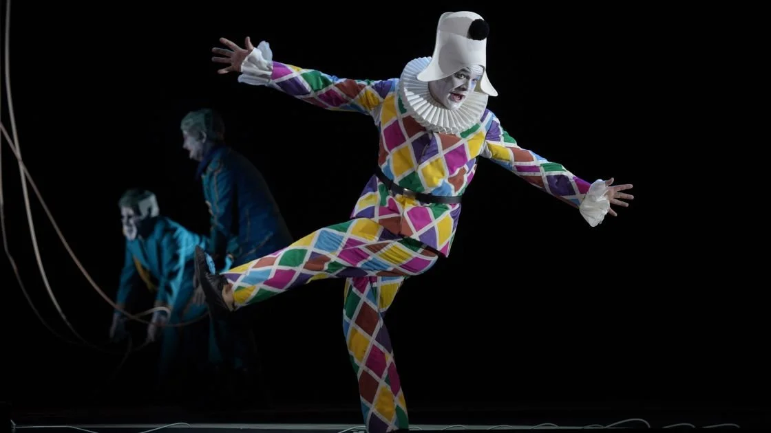 Ein Clown in buntem Kostüm mit Harlekin-Muster tanzt oder performt, während im Hintergrund zwei weitere Personen im blauen Kostüm erscheinen, alle vor einem schwarzen Hintergrund.