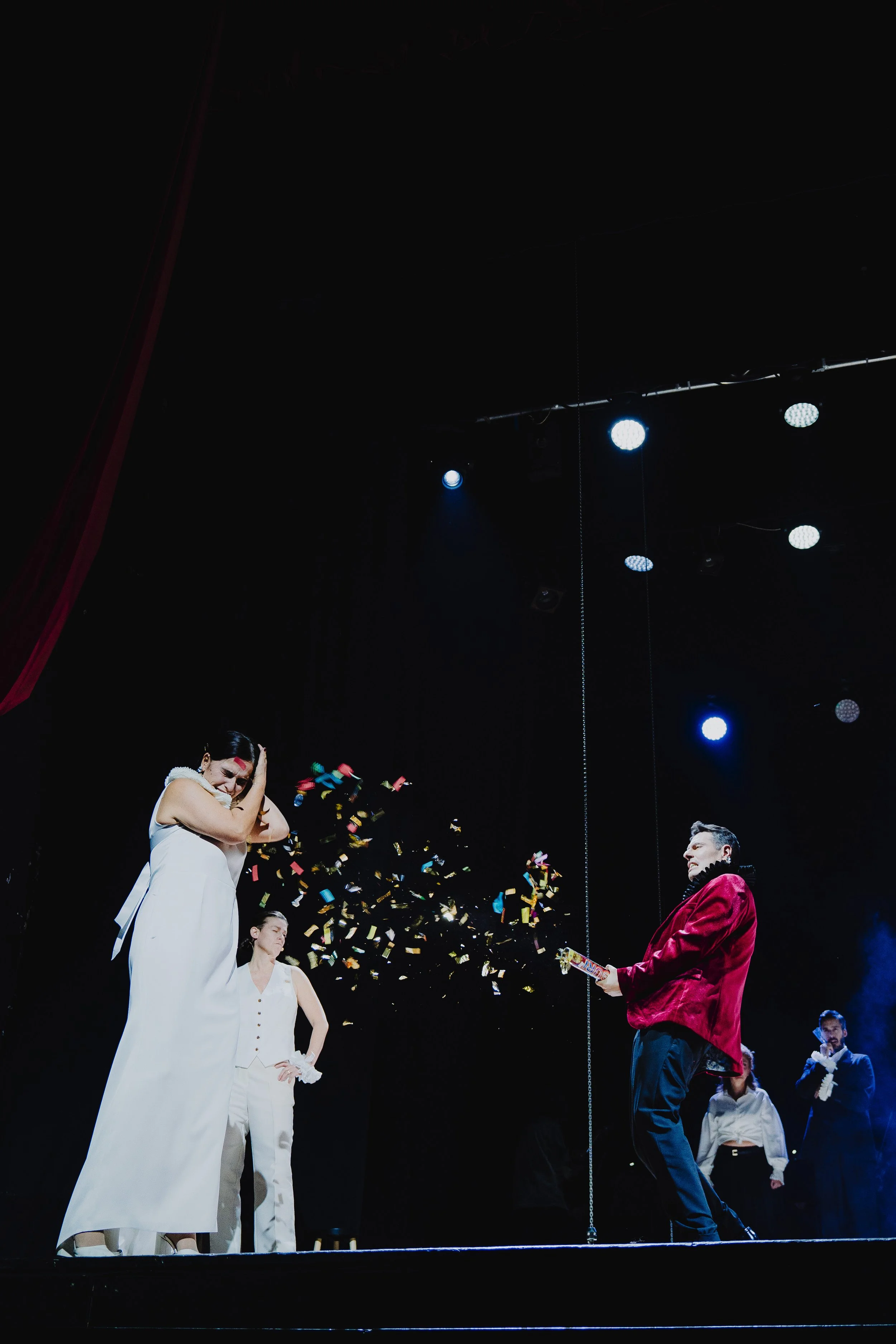 Bühnenperformance mit mehreren Menschen, einer Frau in weißem Kleid, die sich die Ohren zuhält, und einem Mann in rotem Jackett, der von einem Knallkörper Feuerwerk bekommt, Konfetti fliegt durch die Luft.
