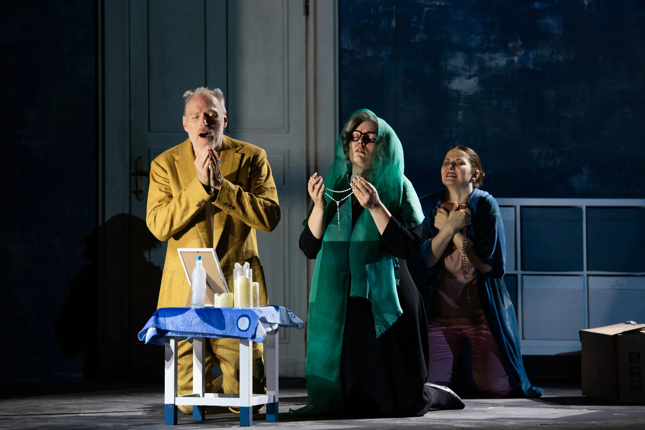 Drei Menschen in einer religiösen Szene, einer mit verschränkten Händen, einer in einem grünen Schleier, die andere kniet auch mit einem Kreuz in der Hand, auf einem Bühnenbild mit Kerzen und einem Tisch.