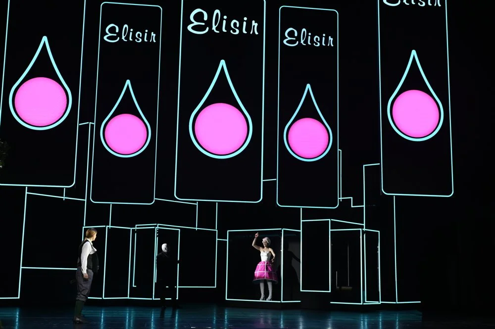Bühnenperformance mit neonartigen Linien und großen Schildern mit der Aufschrift 'Elisin' sowie pinken Tropfen, zwei Personen auf der Bühne, eine in einem pinken Kleid und die andere in dunkler Kleidung.
