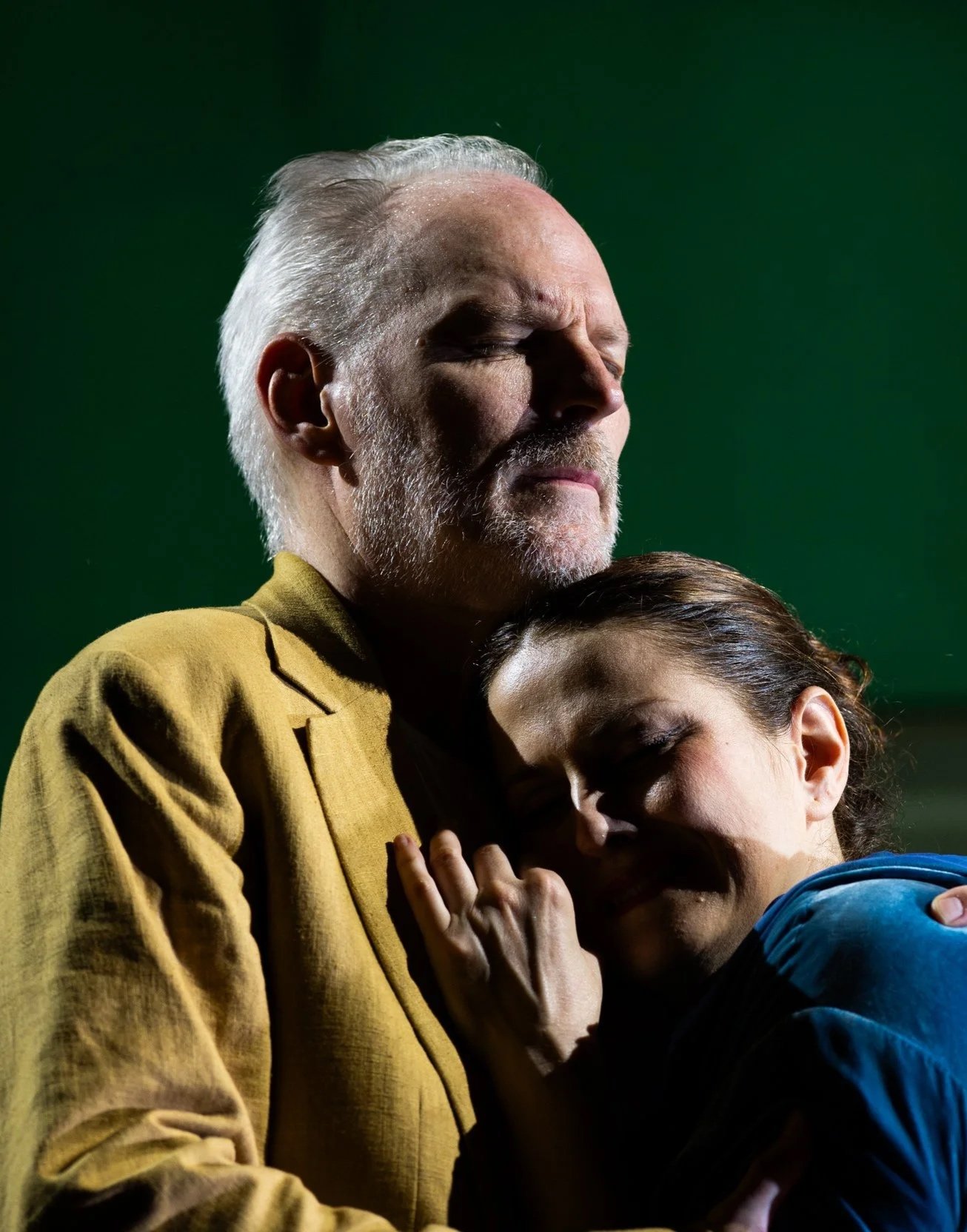 Ein Mann mit grauem Haar und Bart umarmt eine Frau, beide mit geschlossenen Augen, in einer herzlichen Umarmung vor einem grünen Hintergrund.