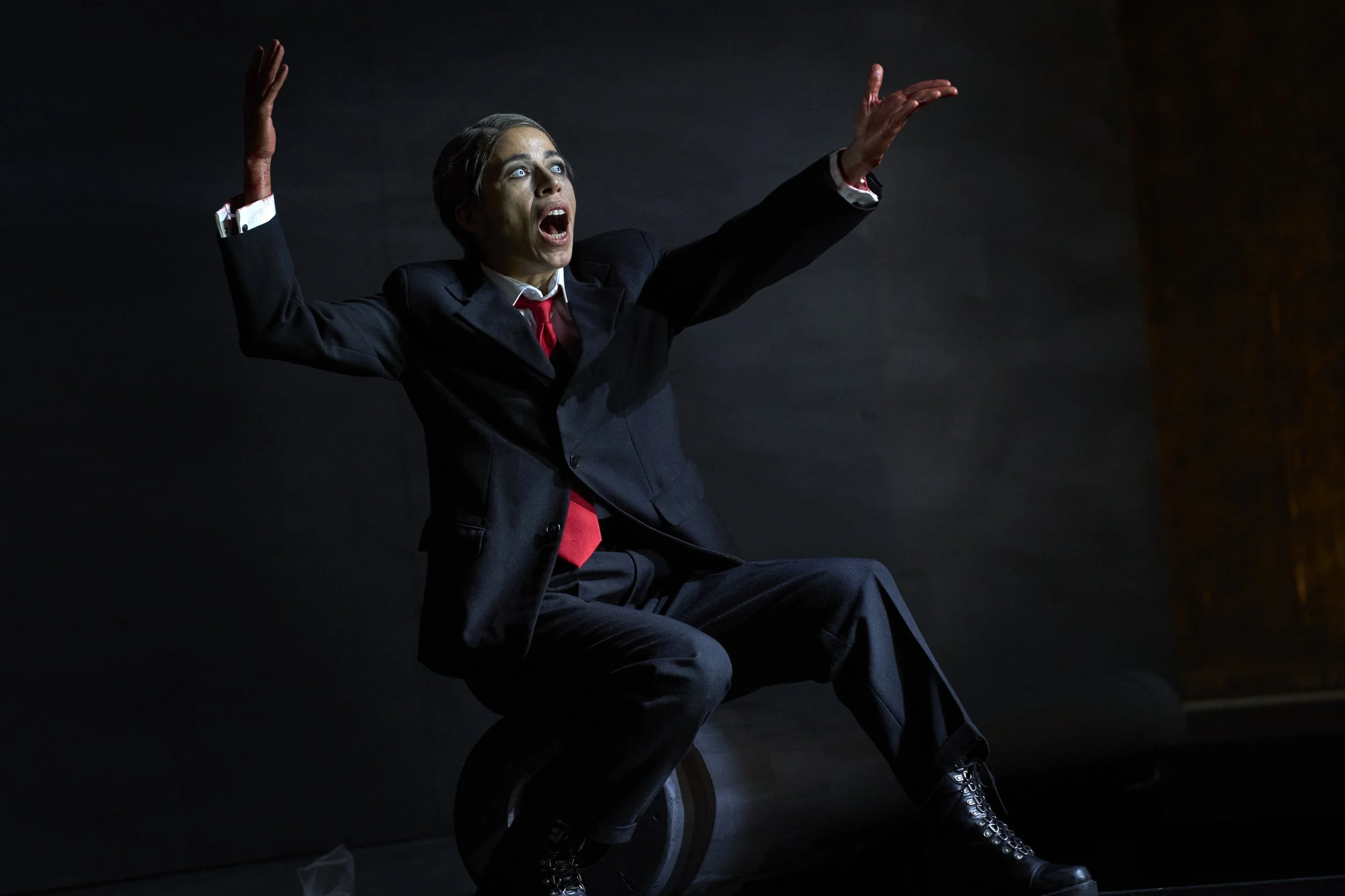 Eine Person in formeller Kleidung sitzt auf einem unebenen Objekt, mit erhobenen Armen und einem Ausdruck von Schock oder Angst, vor einem dunklen Hintergrund.