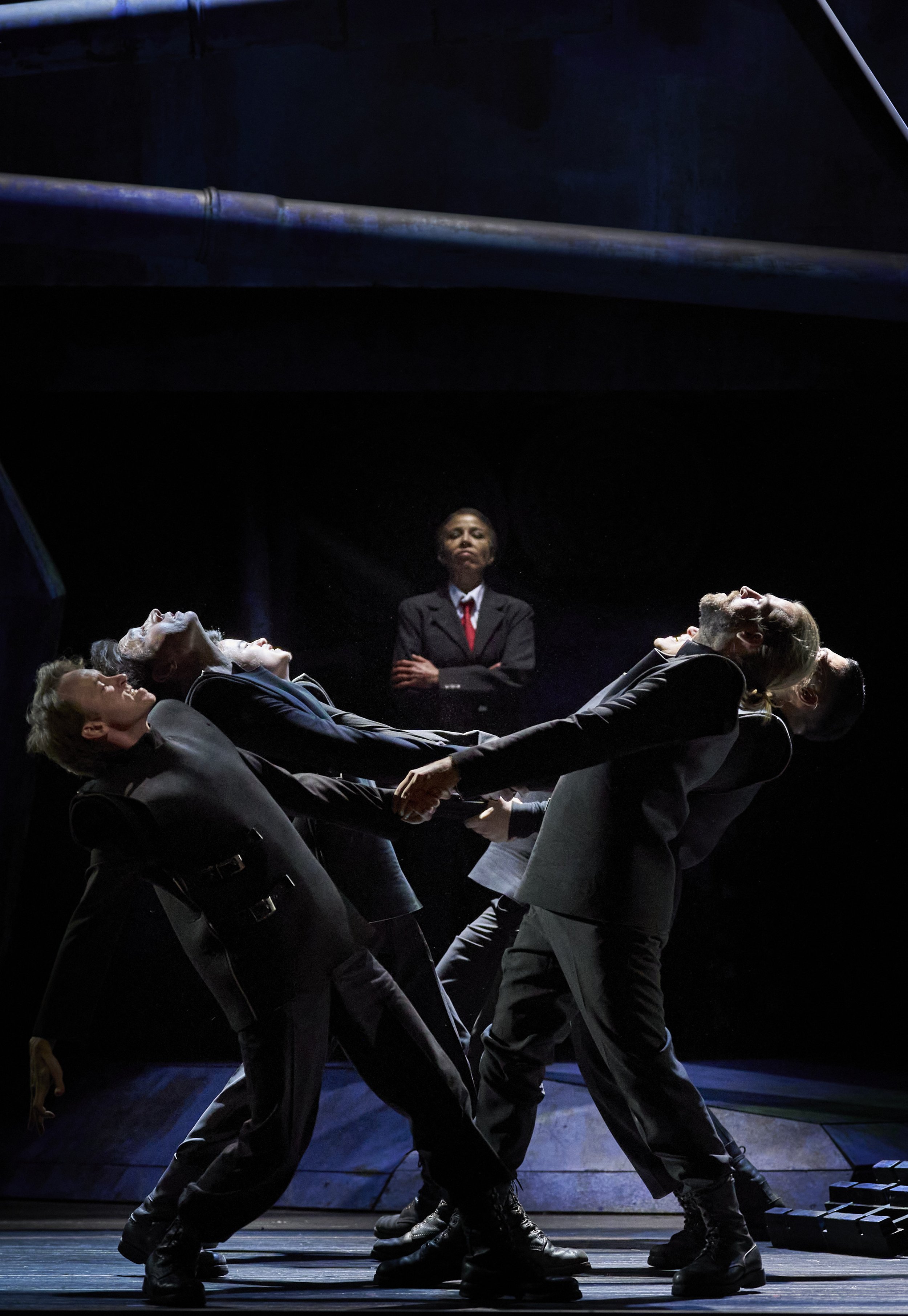 Theaterstück mit sechs Schauspielern auf der Bühne, fünf Männer, die sich an den Händen halten und in einer Gruppe eine emotionale Szene darstellen, während eine Frau im Anzug im Hintergrund zuschaut mit verschränkten Armen.