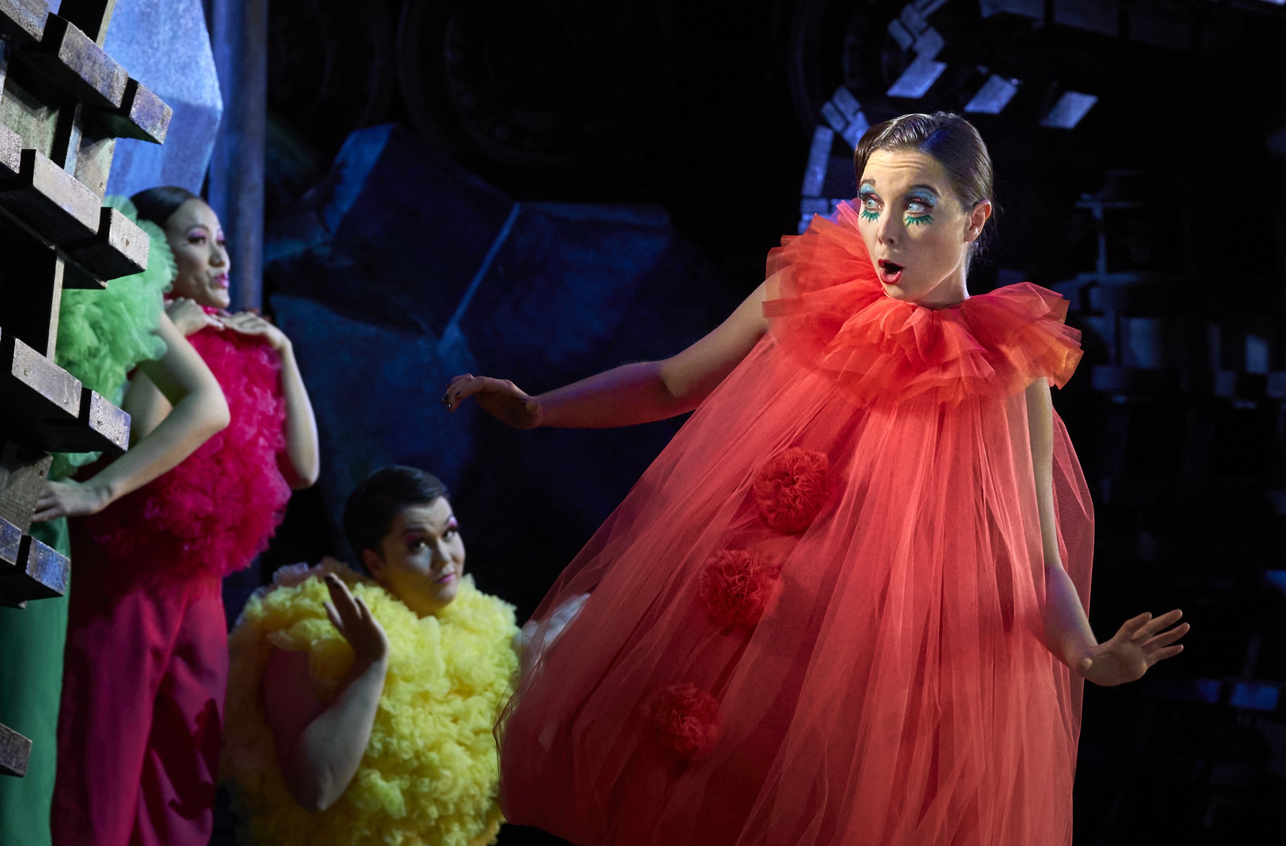 Menschen in bunten, extravaganten Kostümen bei einer Theateraufführung, eine Frau in einer roten Tüllkleidung im Vordergrund, die andere in grüner, pinker und gelber Kleidung im Hintergrund, dunkler Bühnenhintergrund.