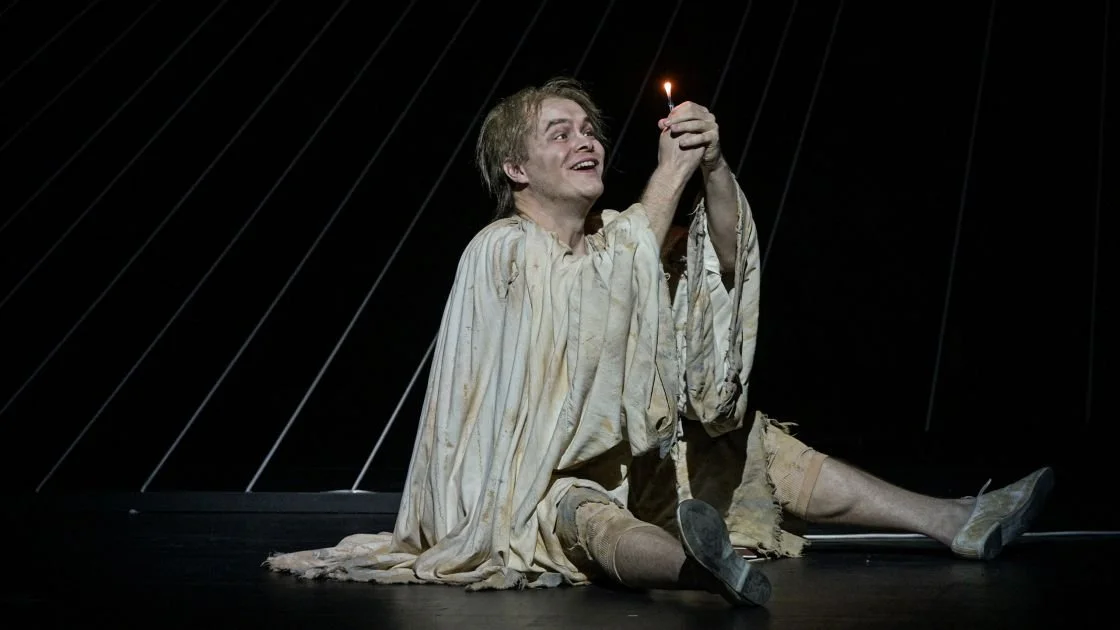 Mann in zerissener Kleidung hält eine kleine Flamme und lächelt auf einer dunklen Bühne.