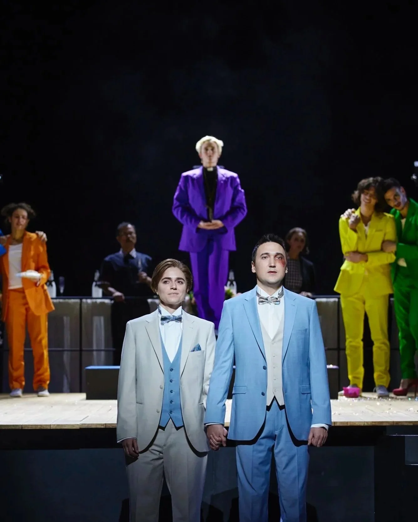 Theatermit Schauspielern in bunten Kostümen auf Bühne, zentrales Paar in weißen Anzügen, umgeben von anderen Figuren in bunten Outfits, eine Person in lila auf Podest im Hintergrund.