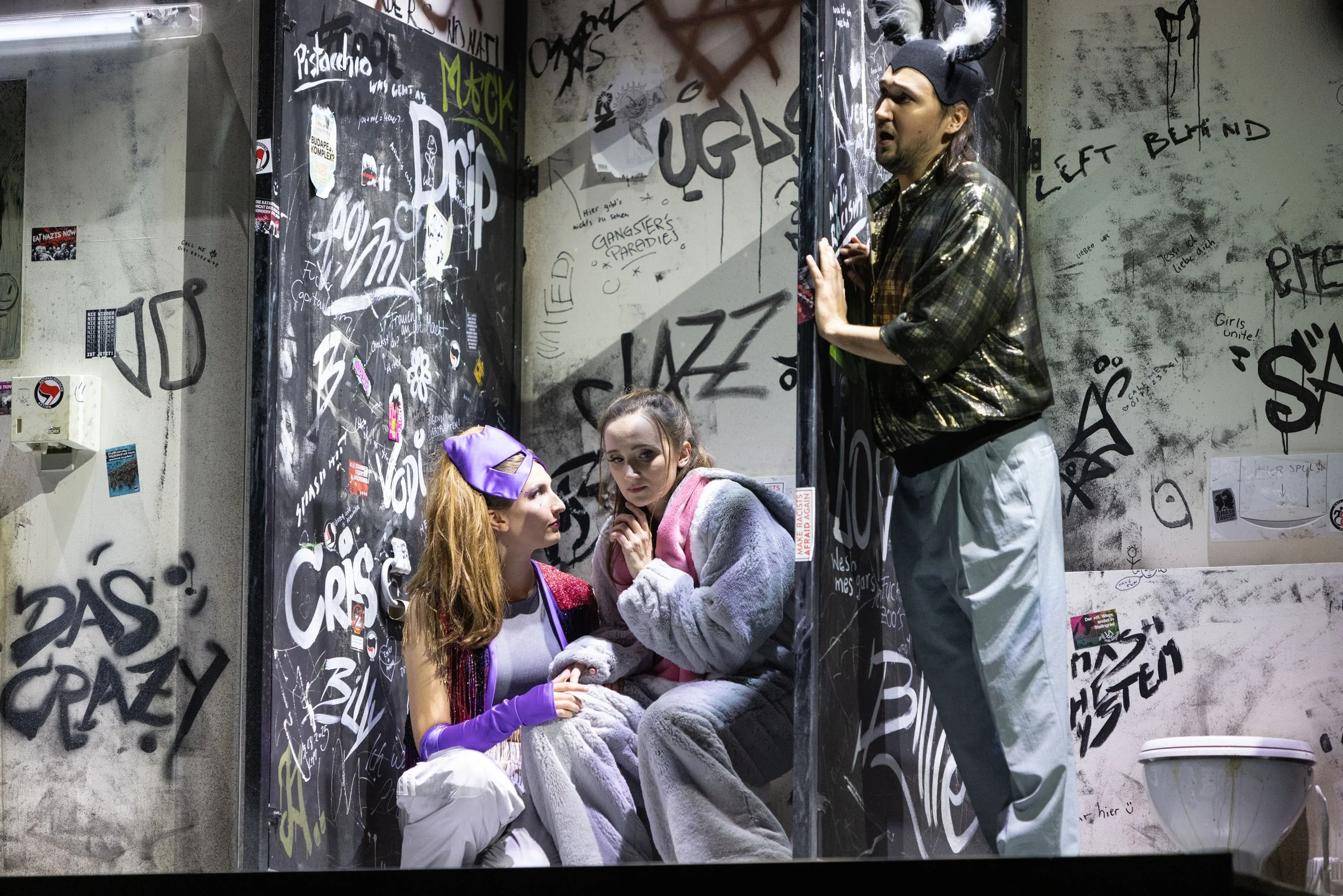 Drei Menschen in einer grauen, graffitiüberwachsenen Toilette. Zwei Frauen sitzen auf dem Boden, eine trägt eine lilafarbene Maske und Handschuhe, die andere trägt einen verspielt grauen Overall. Der Mann steht vor ihnen, trägt eine schwarze Mütze mi