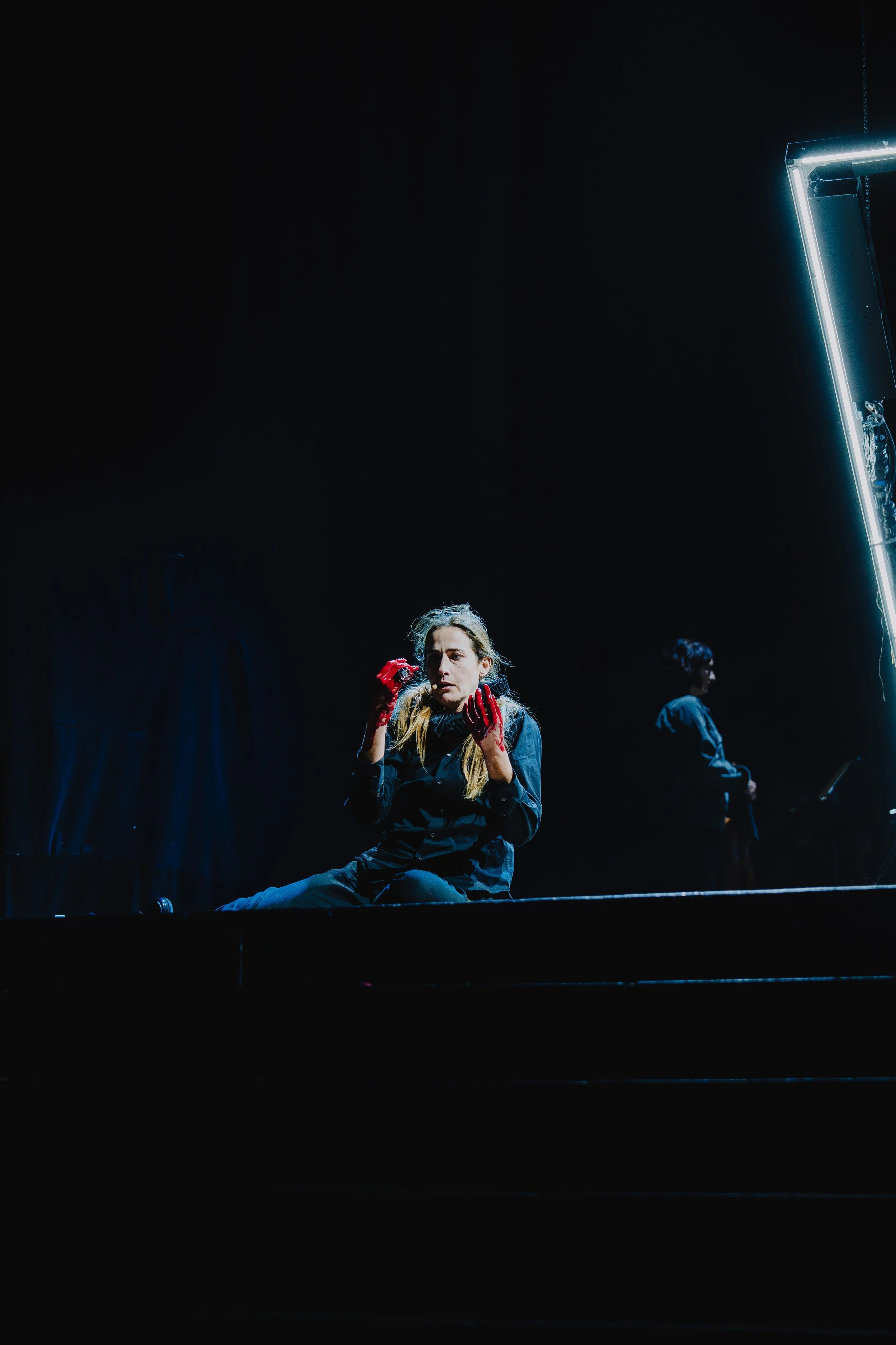 Eine junge Frau sitzt auf dem Boden, trägt schwarze Kleidung und rote Handschuhe, wirkt nachdenklich, im Hintergrund eine weitere Person steht in einem dunklen Raum mit Neonbeleuchtung.