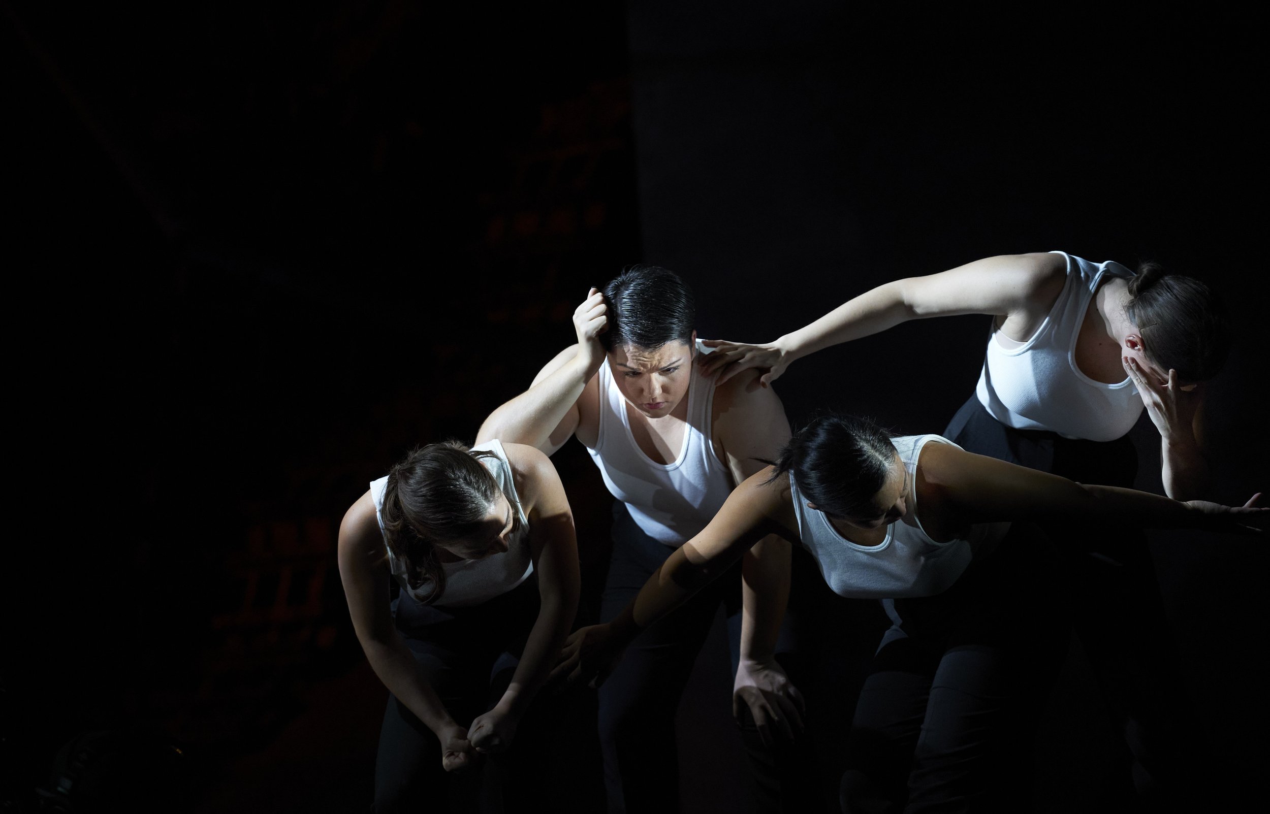 Vier Frauen in weißen T-Shirts und schwarzen Hosen in einem dunklen Raum, die eine dramatische Pose einnehmen, mit unterschiedlichen Gesichtsausdrücken und Gesten.