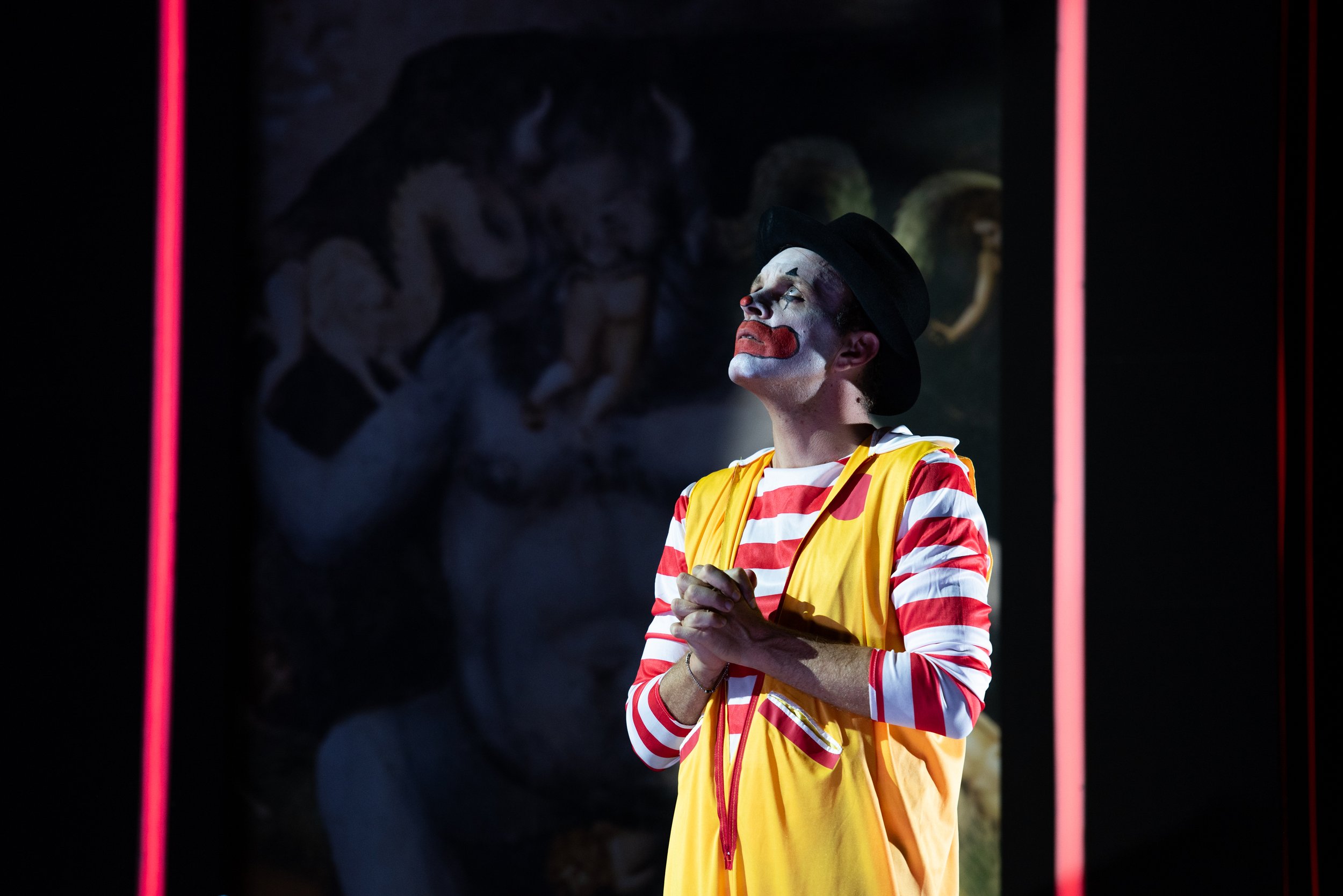 Clown mit weißen Gesichtsmakeup, rotem Lippenstift, rotem und weiß gestreiftem Hemd, gelber Weste und schwarzem Hut steht vor einem dunklen Hintergrund mit einem Gemälde im Schatten.