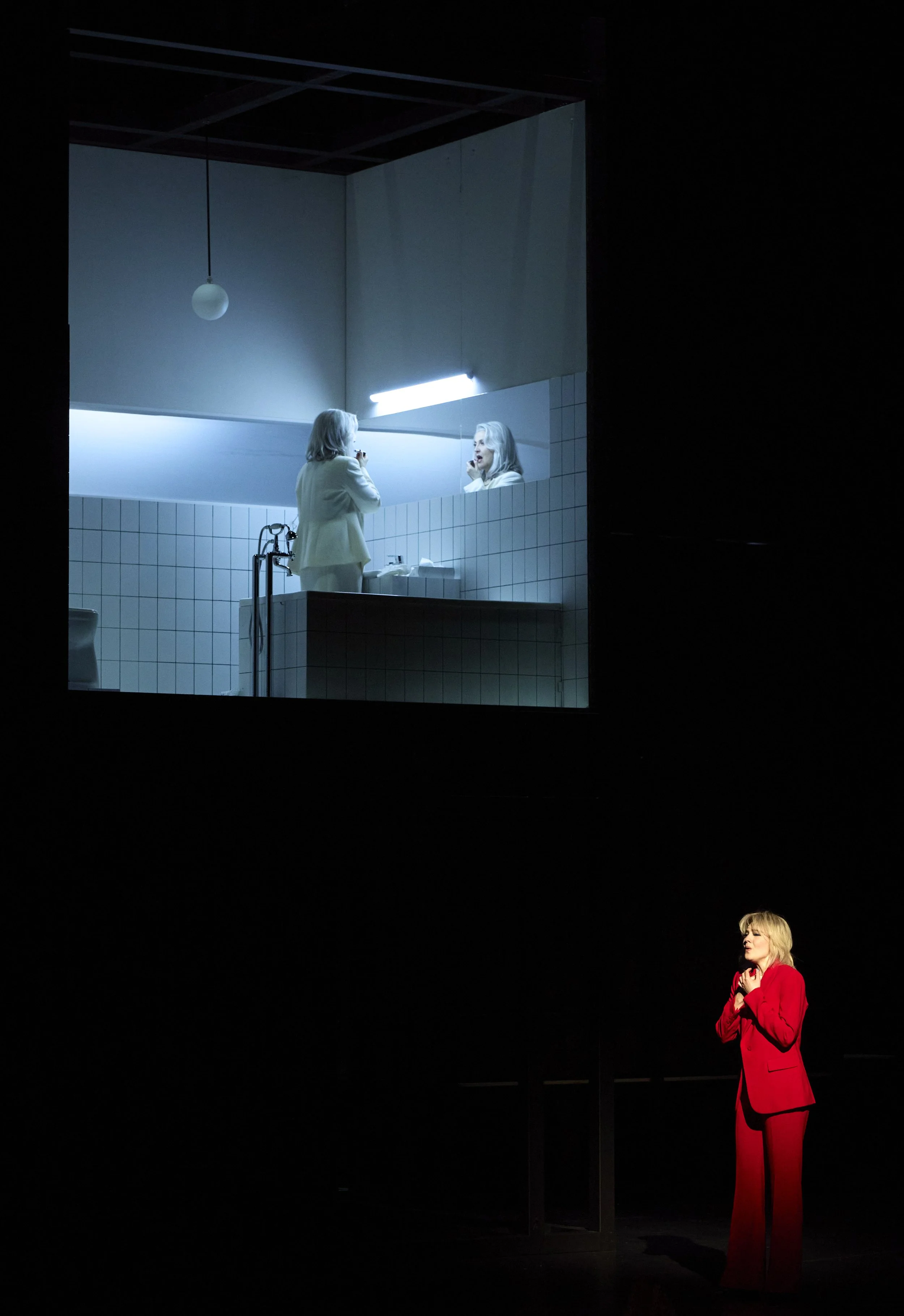 Auf der Bühne eine Frau in rotem Anzug, die in Gedanken versunken wirkt, und im Hintergrund ein Projektor, der eine Szene eines Badezimmers zeigt, in dem eine ältere Frau vor einem Spiegel steht.