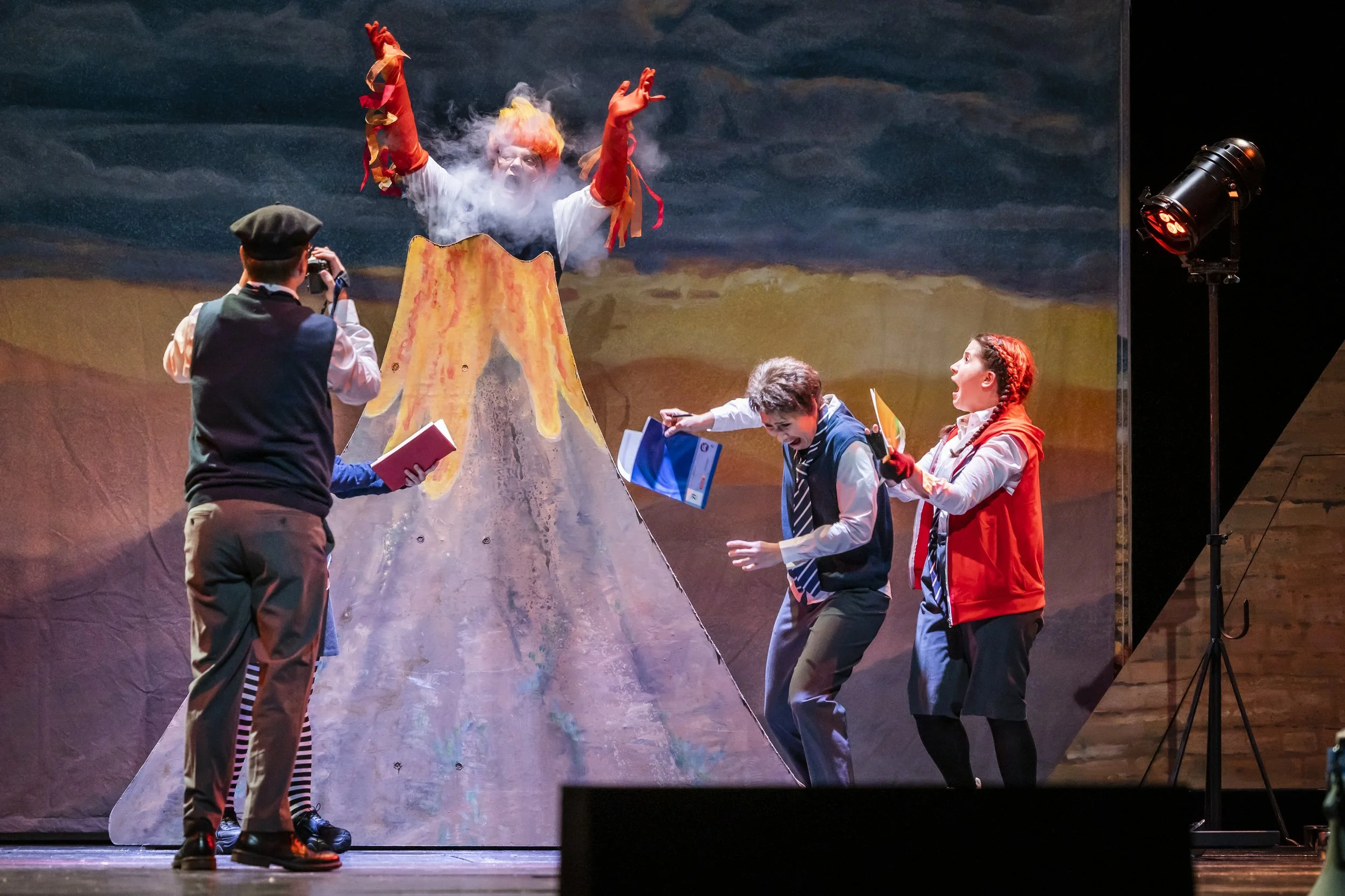 Schauspieler in einem Theaterstück, das einen Vulkan darstellt, mit einem Schauspieler, der Rauch aus einem Vulkan sprüht, während andere Zuschauer mit Büchern oder Requisiten reagieren.