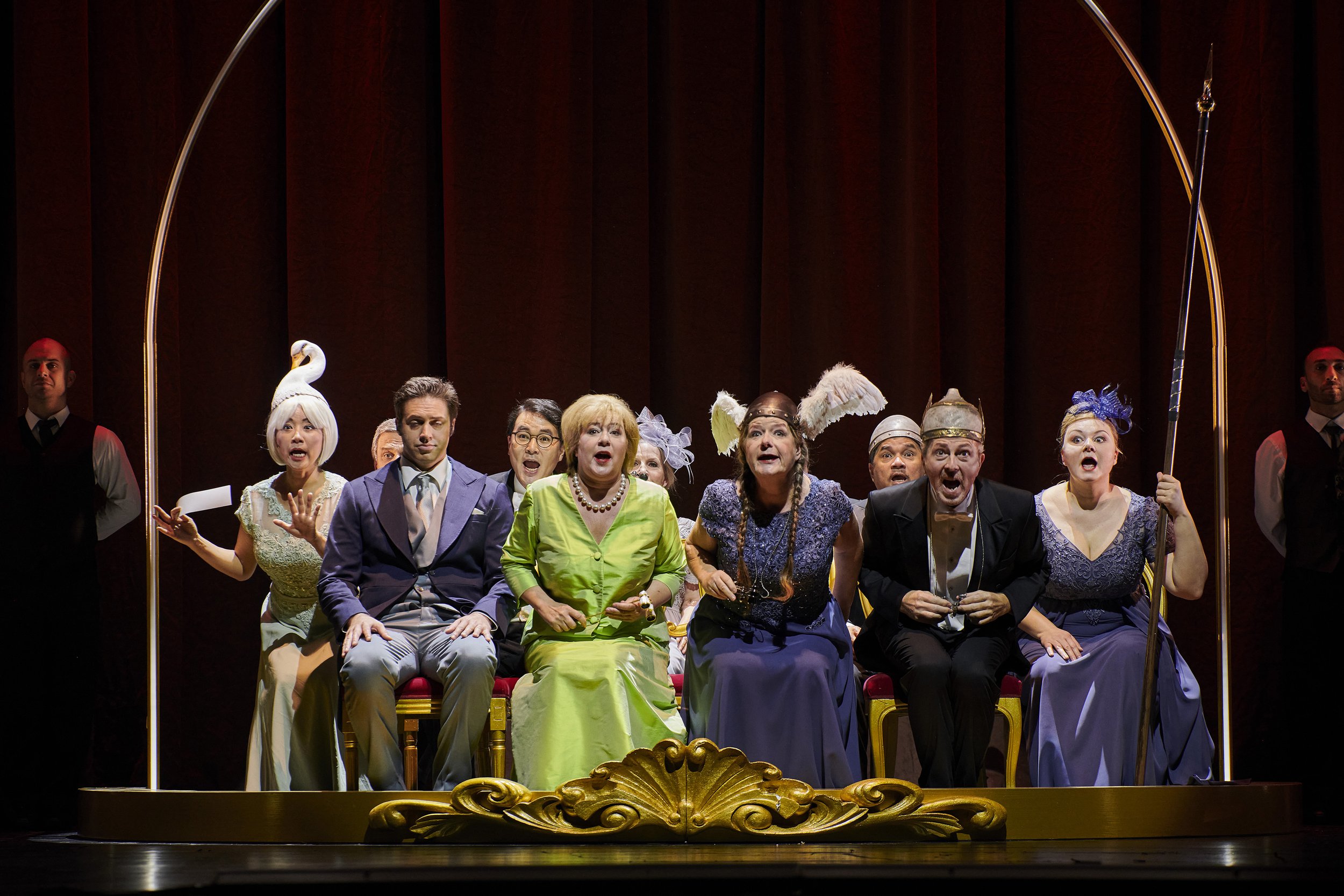 Schauspieler auf einer Bühne in einer Theaterproduktion, sitzend in einem Marquis, verkleidet mit bunten, auffälligen Kostümen, mit Panik und Überraschung im Gesicht, umgeben von Zuschauern und Bühnenhintergrund.