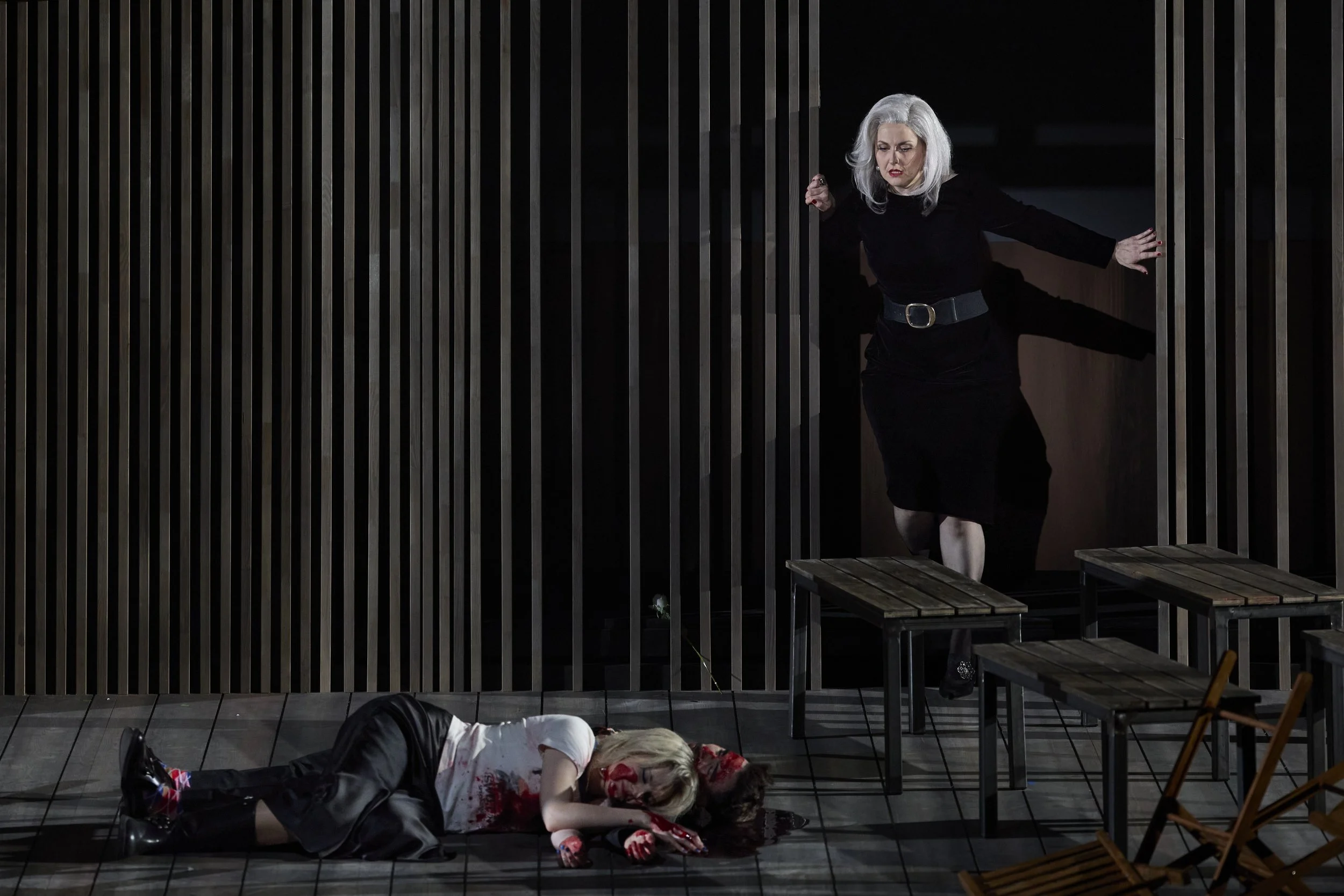 Eine Szene aus einem Theaterstück mit zwei Frauen. Eine Frau liegt blutüberströmt auf dem Boden, die andere Frau steht in der Türöffnung und schaut entsetzt.