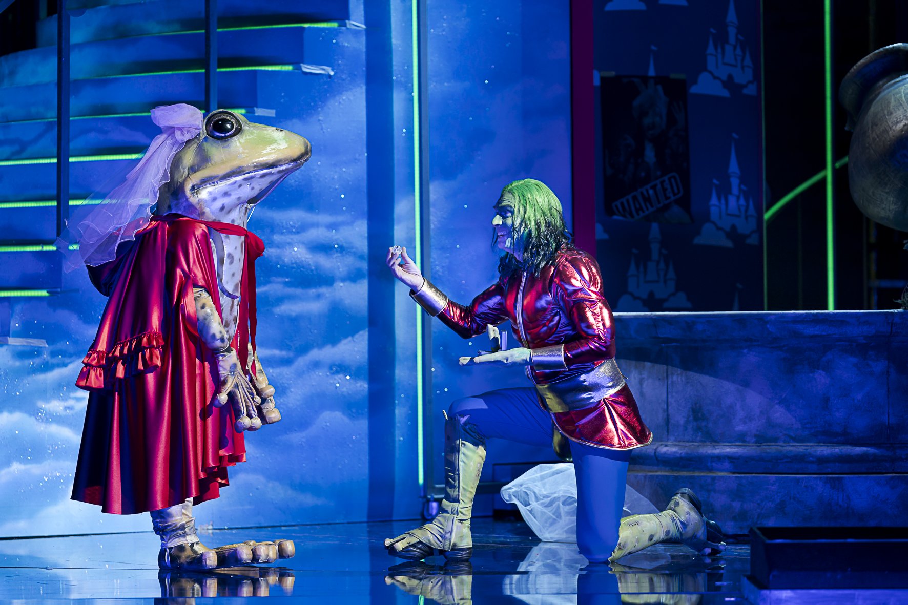 Ein Mensch im Joker-Kostüm kniet auf einem Boden und spricht mit einer anthropomorphen Froschfigur, die ein rotes Kleid und einen Schleier trägt, alles vor einem bunten Bühnenhintergrund.