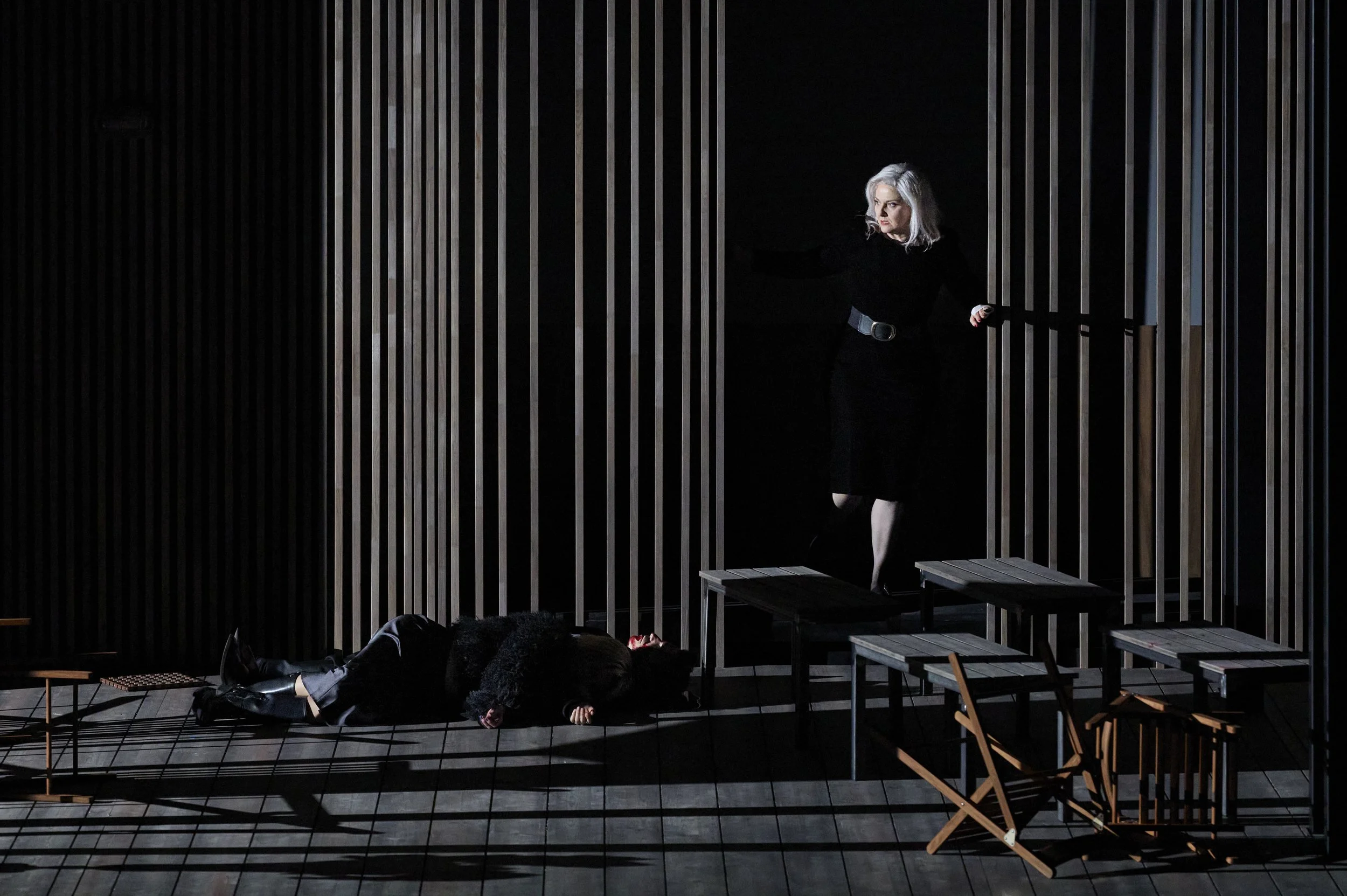 Szene eines Theaterspiels mit einer Frau an einem Geländer, die einen anderen Menschen auf dem Boden ansieht, bei eingeschränkter Beleuchtung.