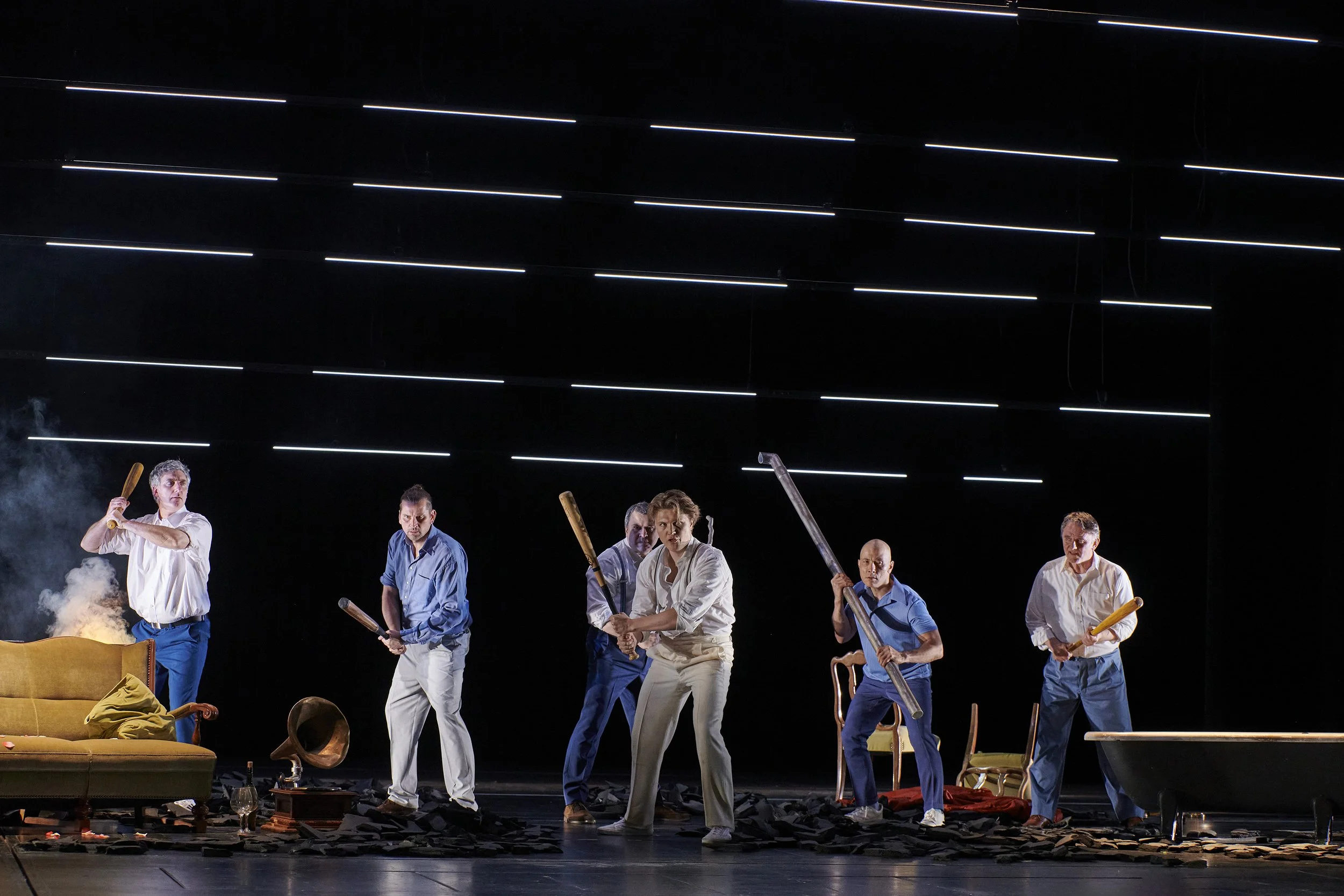 Bühnenbild mit sechs Schauspielern, die mit Gegenständen wie Holzknüppeln und Sägen agieren, in einem dunklen Raum mit moderner Lichtinstallation an der Decke.