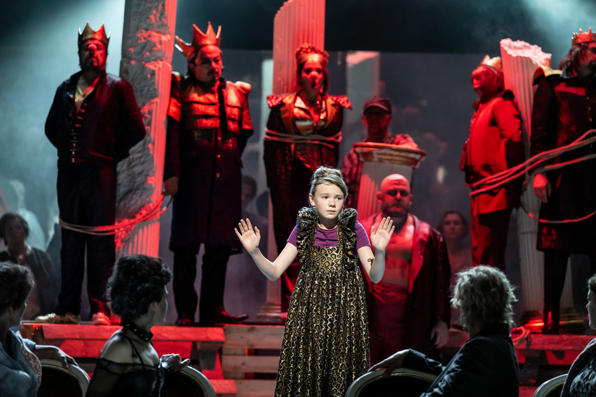 Eine junge Mädchen steht auf einer Bühne, umgeben von Menschen und Schauspielern in Kostümen, die eine Szene in einem Theaterstück darstellen.