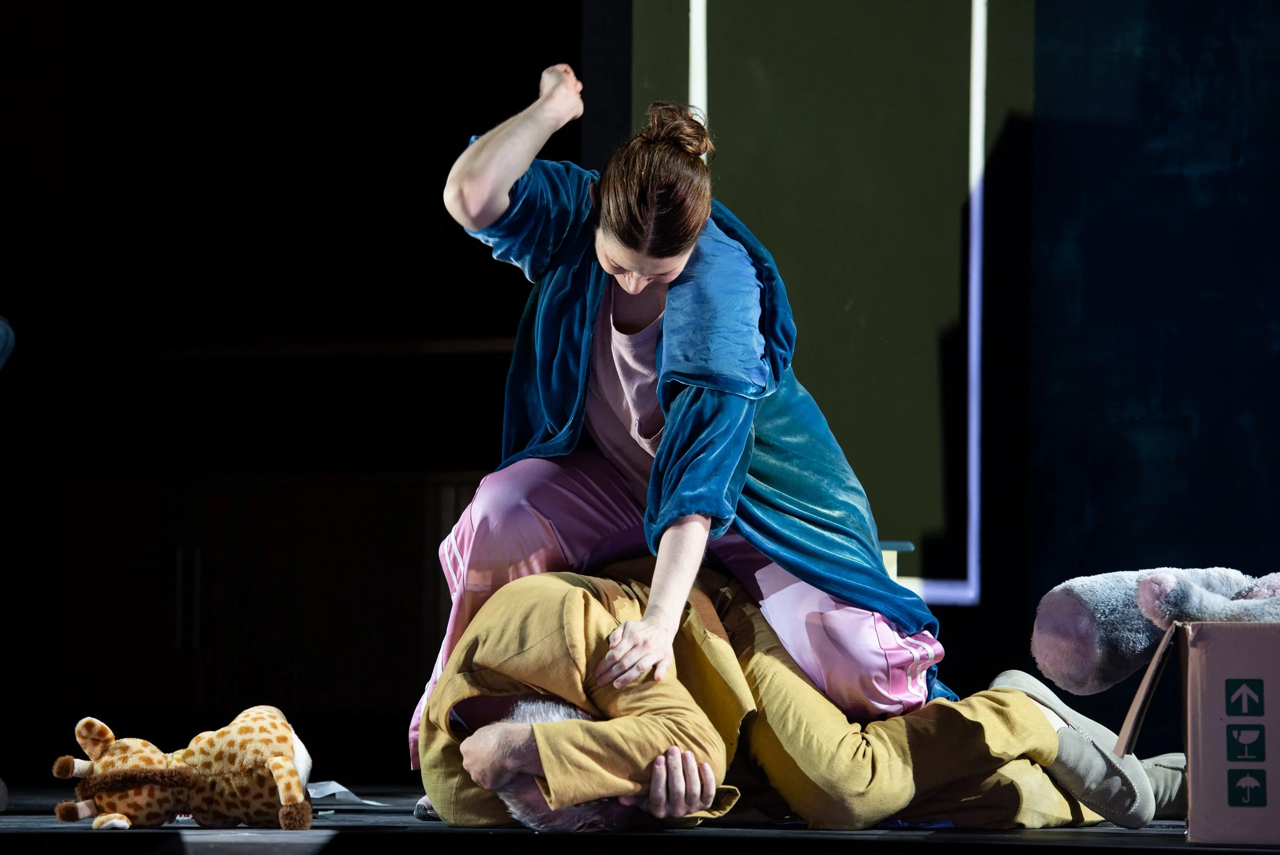 Zwei Frauen bei einer theatralischen Szene, eine sitzt auf dem Boden, die andere steht über ihr und greift sie an, um sie zu schlagen. Es gibt teddyartige Stofftiere und eine Box im Vordergrund.