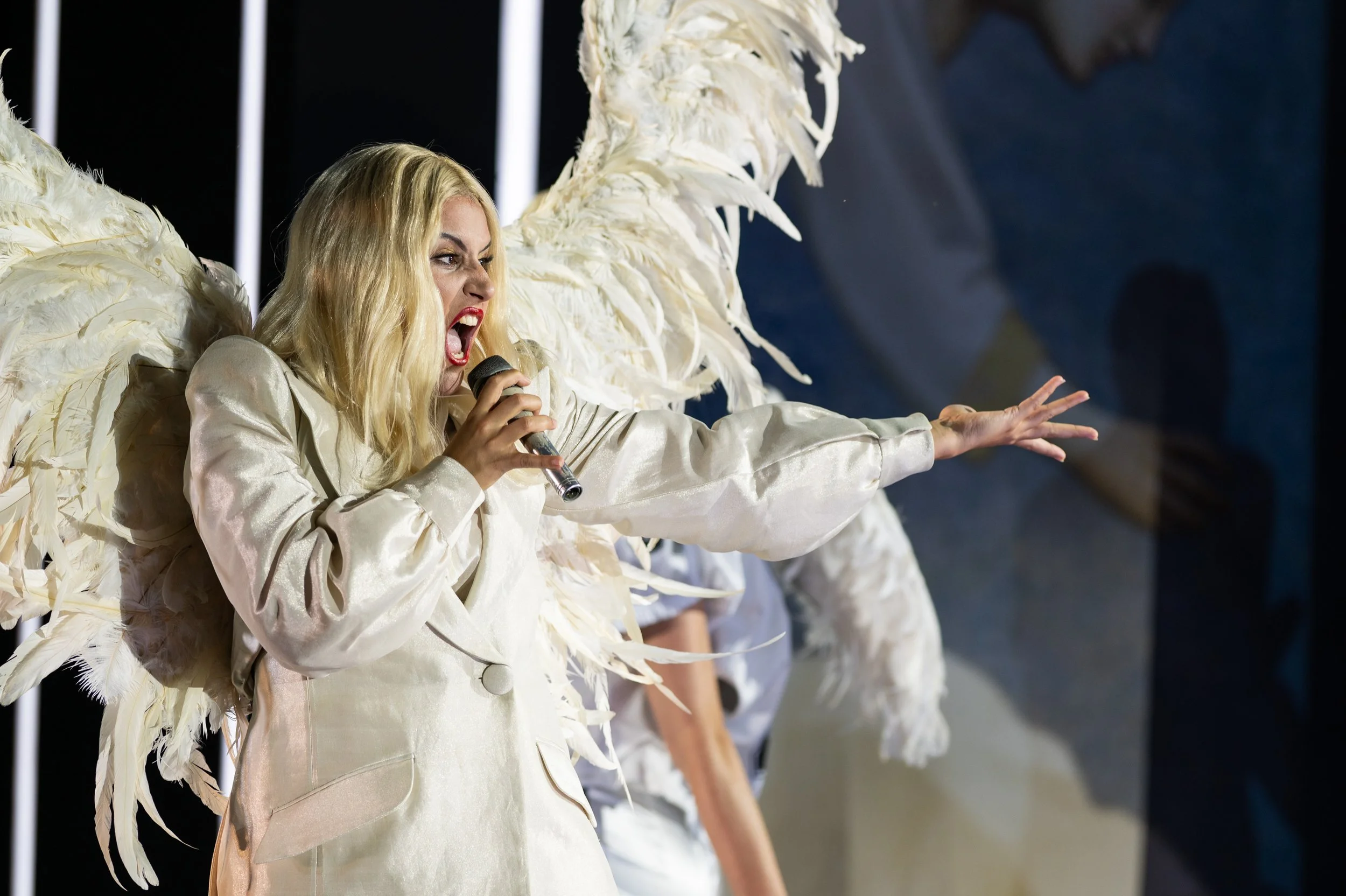 Eine blonde Frau in einem weißen Outfit mit großen Flügeln, die singt oder spricht und einen Mikrofon hält, auf einer Bühnenutzung.