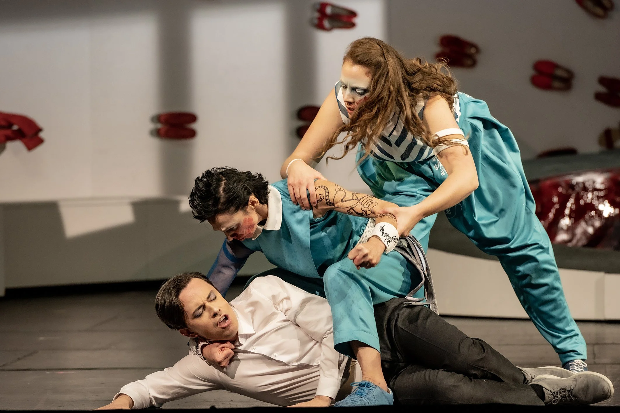 Schauspieler in einer Szene, in der sie sich kämpfend auf dem Boden befinden, mit einer betroffenen Person im weißen Hemd und dunklen Hosen, die am Boden liegt, während zwei andere Körper die Szene bestimmen.