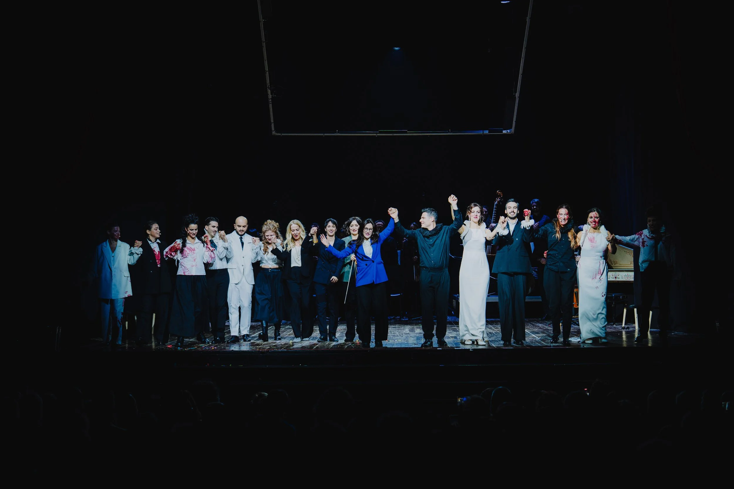 Eine Gruppe von Menschen steht auf einer Bühne, hält sich an den Händen und verbeugt sich nach einer Aufführung. Es sind Männer und Frauen in formeller Kleidung, einige in dunklen Anzügen und Anzügen, andere in weißen Kleidern, einige haben rote Farb