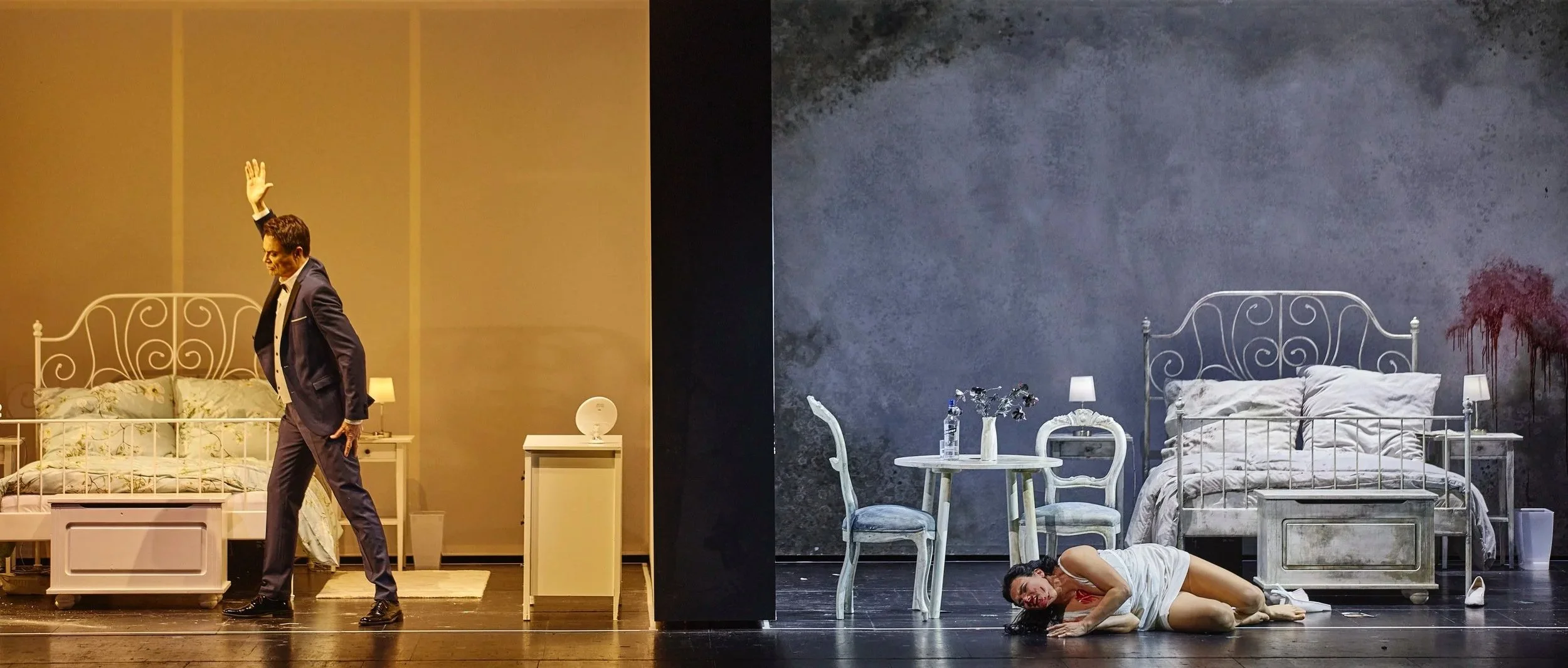 Bühnenbild mit zwei getrennten Szenen: links ein Mann in Anzug steht vor einem Bett, rechts eine Frau liegt auf dem Boden neben einem Bett, im Hintergrund ein dunkler Wandabschnitt, mit einem Tisch, Stühlen und Dekorationsgegenständen.