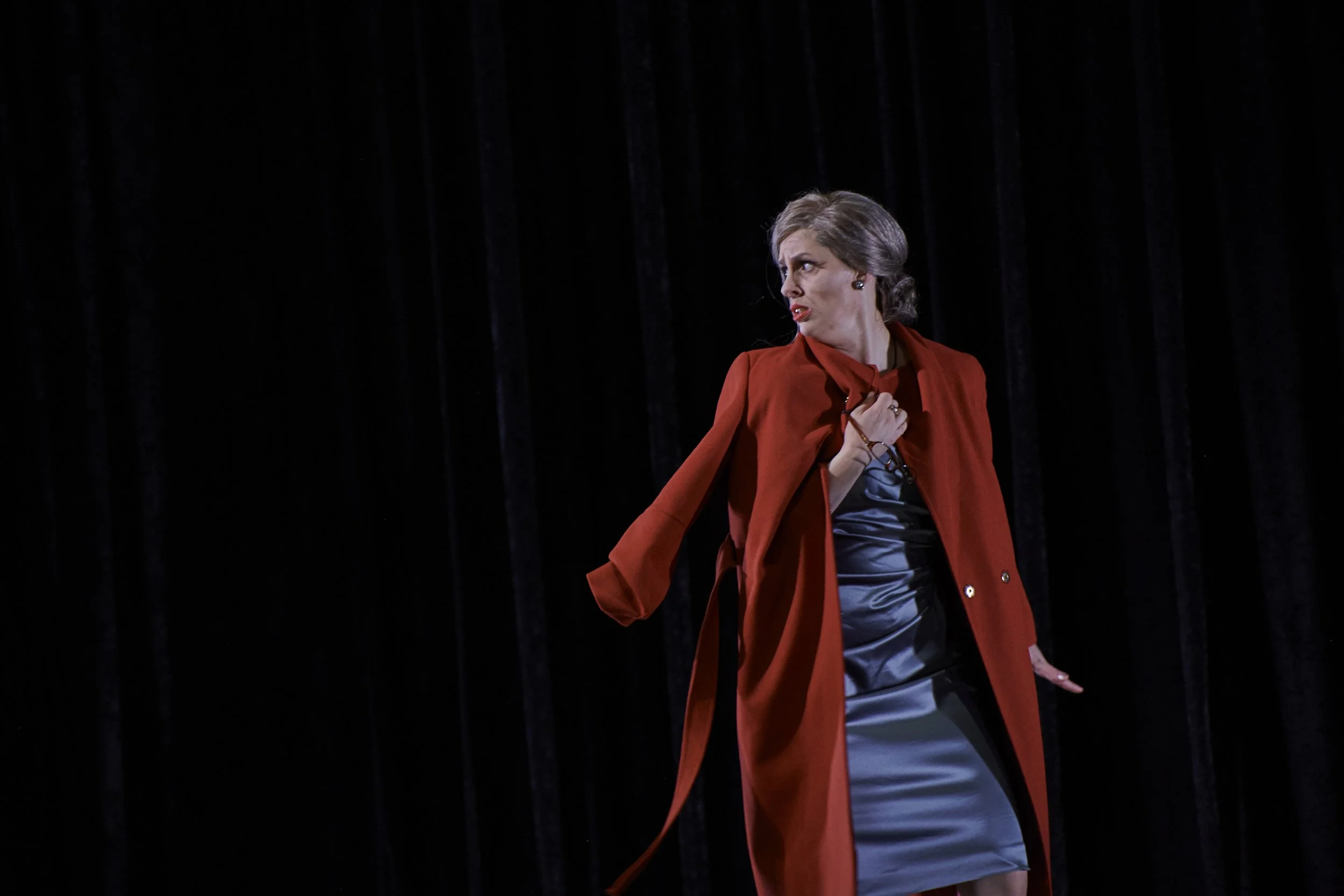 Schauspielerin auf Bühne in rotem Mantel, zeigt ausdrucksstarke Geste, dunkler Hintergrund.