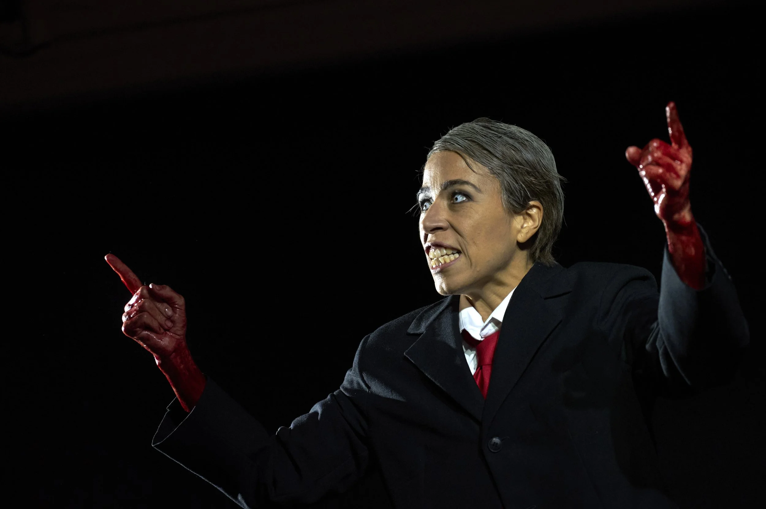 Frau in dunklem Anzug mit blauen Augen und kurzen grauen Haaren, die mit erhobener Hand und roten, blutenden Fingern in einer aggressiven Haltung ist.