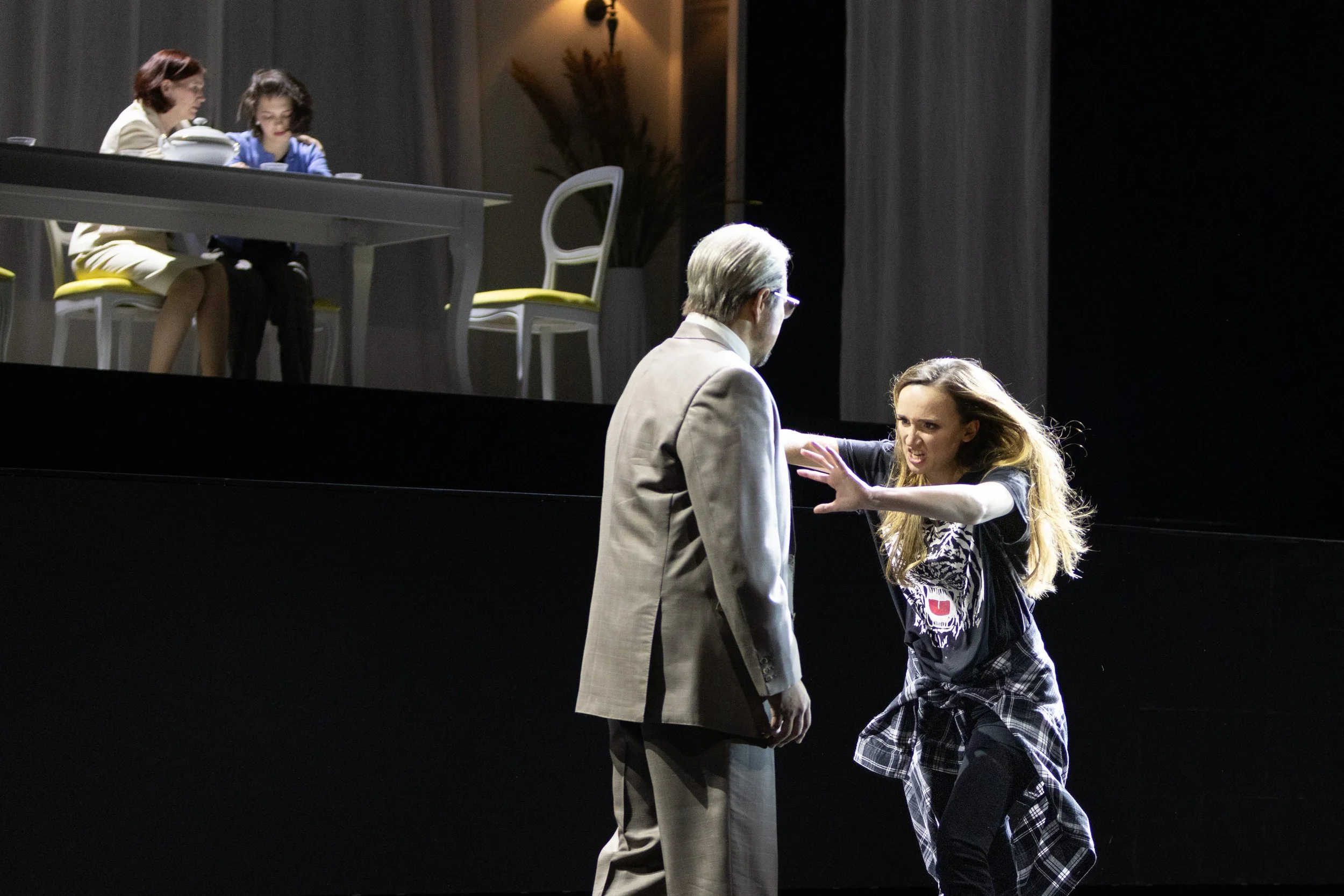 Theater Szene mit einem älteren Mann in Anzug und einer jungen Frau, die sich streiten, im Hintergrund ein Tisch mit zwei Frauen, die lesen oder telefonieren.