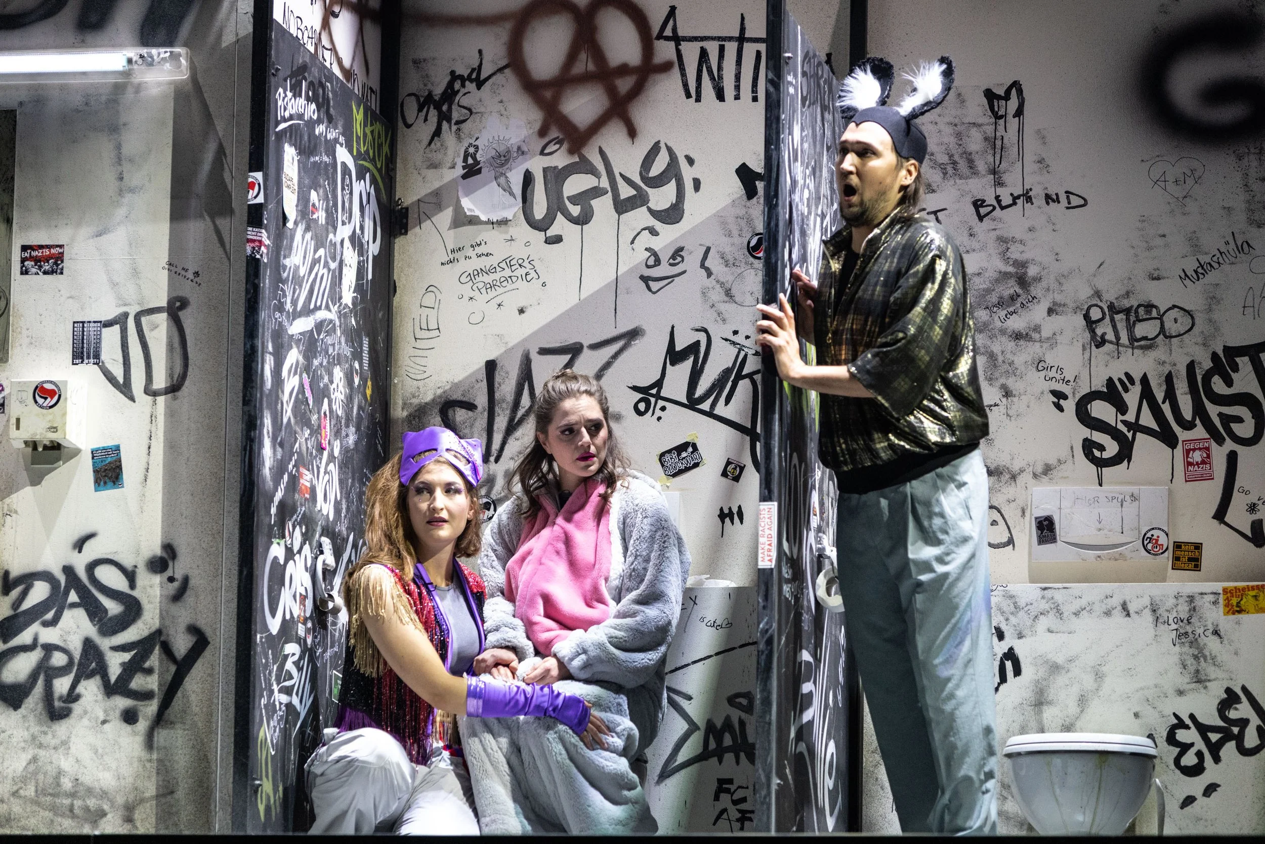 Eine Szene im Waschraum mit zwei Frauen, die in einer Kabine sitzen, und einem Mann, der draußen schreit. Der Raum ist mit Graffiti bedeckt.