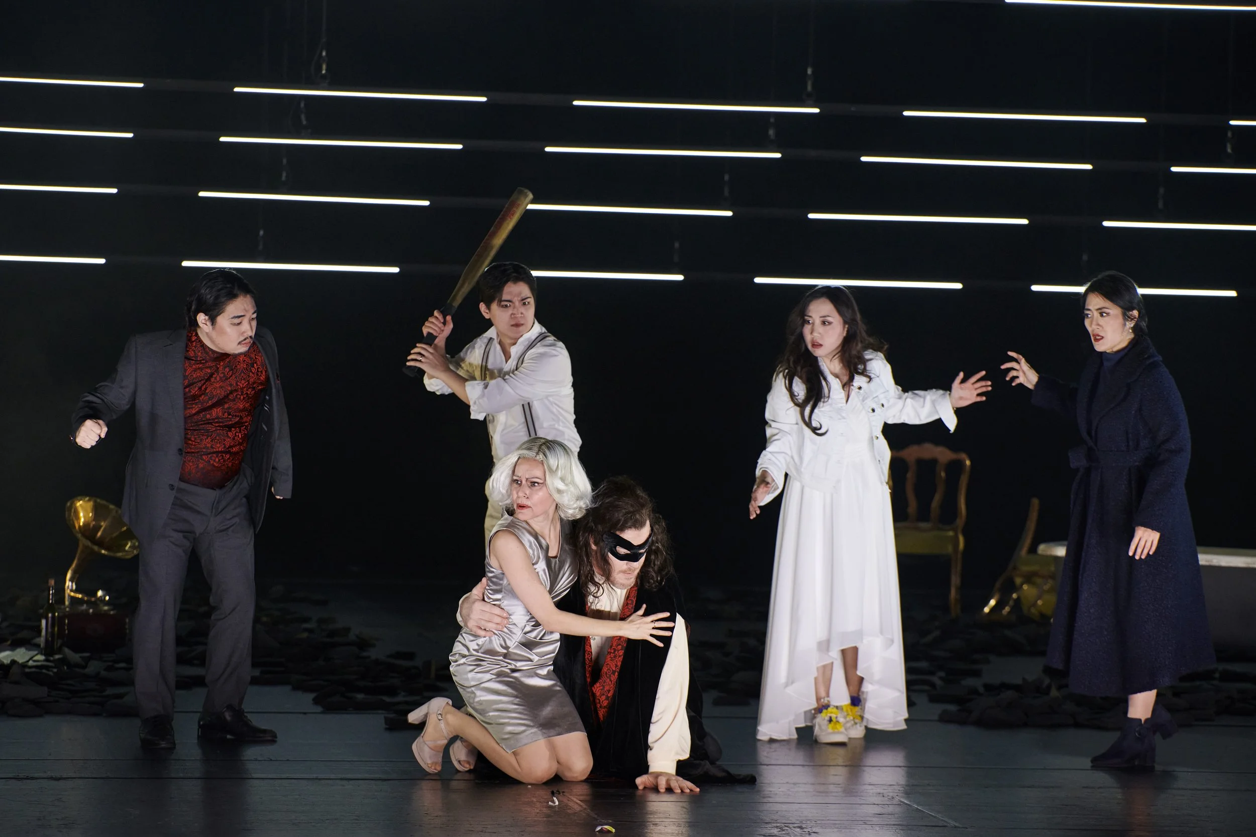 Bühnenaufnahme eines Theatersaustritts mit sieben Schauspielern in unterschiedlichen Posen und Kostümen, im Hintergrund schwarzer Vorhang und moderne Lichtstreifen