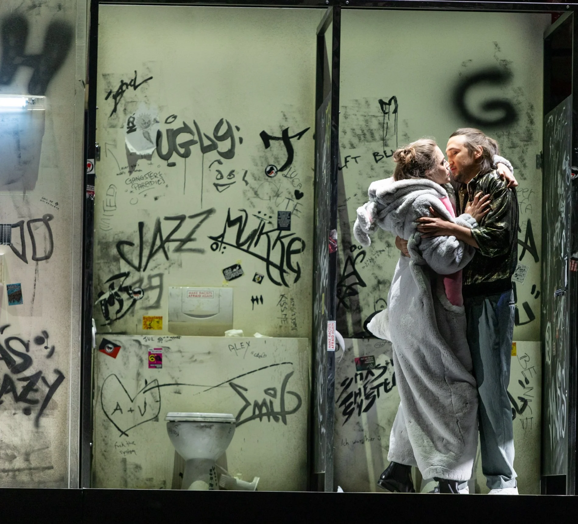 Zwei Menschen, die sich küssen, in einem graffiti-verseuchten öffentlichen Toilettenraum, umgeben von Graffiti und Tags.