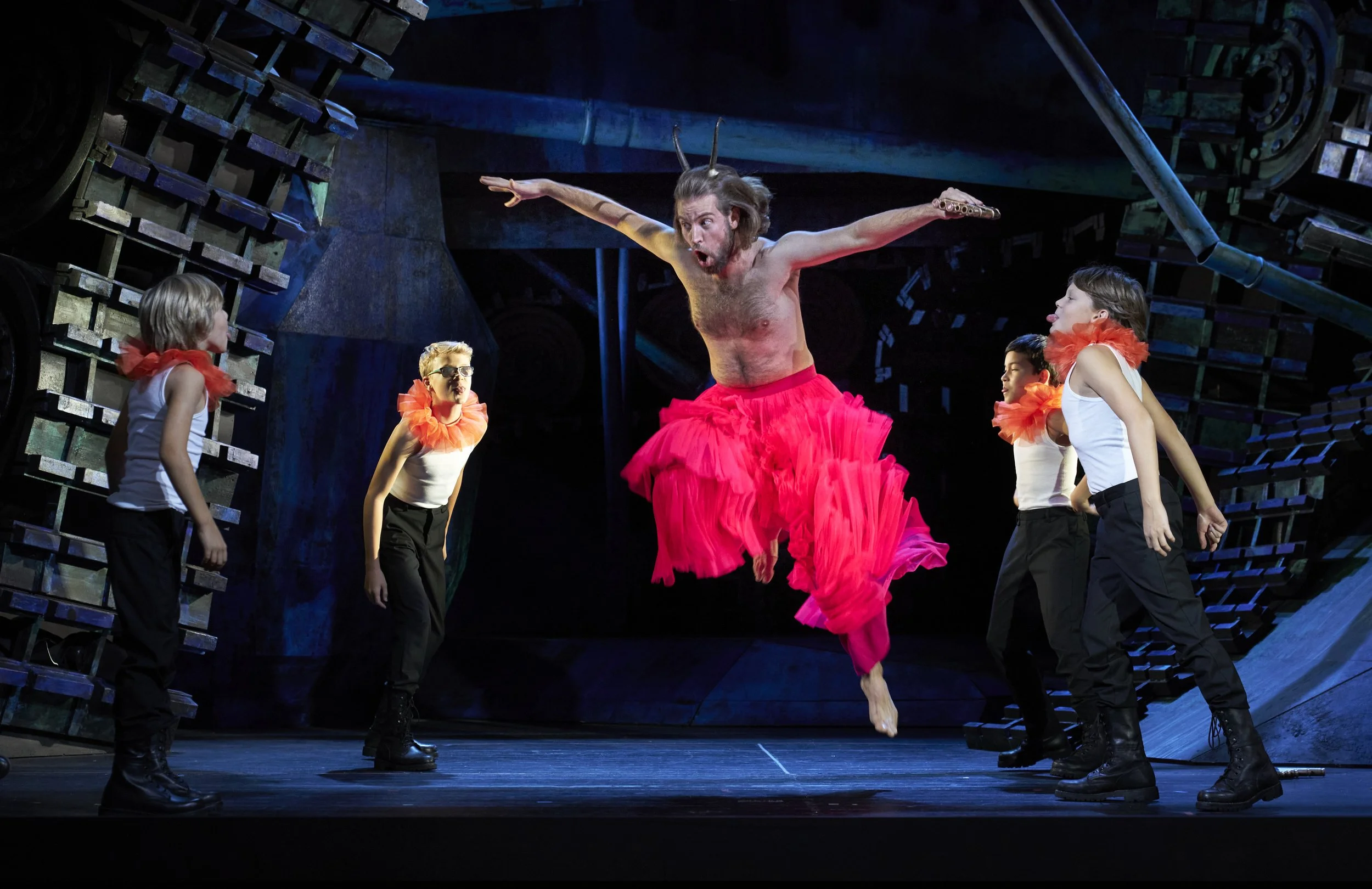 Ein Mann in einem pinken Tüllrock springt auf einer Theaterbühne, umgeben von vier Kindern in weißen Oberteilen, schwarzen Hosen und orange Tüllschleiern, mit einer dunklen, industrialartigen Bühnenkulisse im Hintergrund.