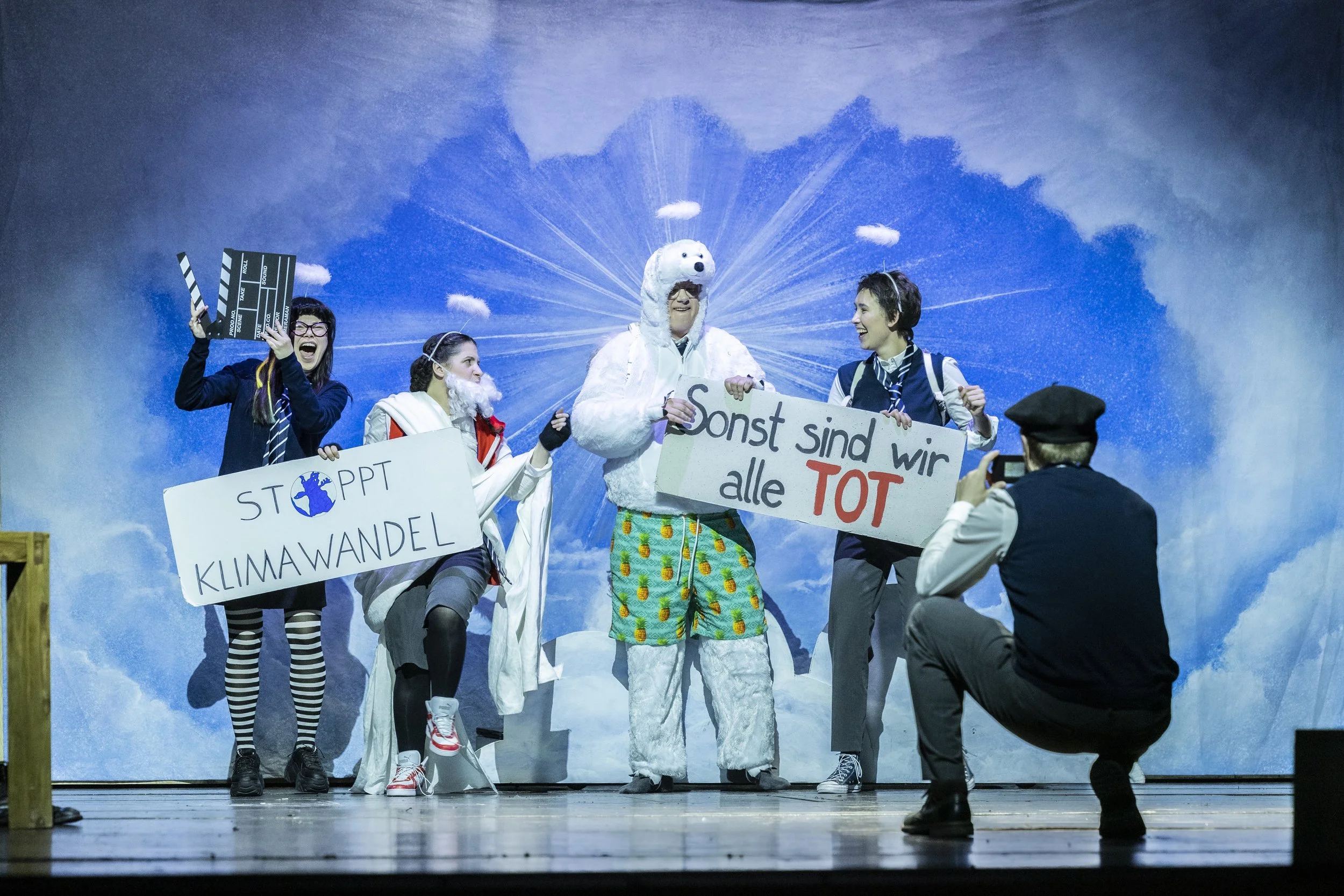 Bühnenszene mit mehreren Menschen in Kostümen, die Klimawandel ansprechen, mit Schildern und einem Mann in einem Polar-Banda Sonnenbrille, das im Mittelpunkt steht, vor einer himmelartigen Hintergrundprojektion.