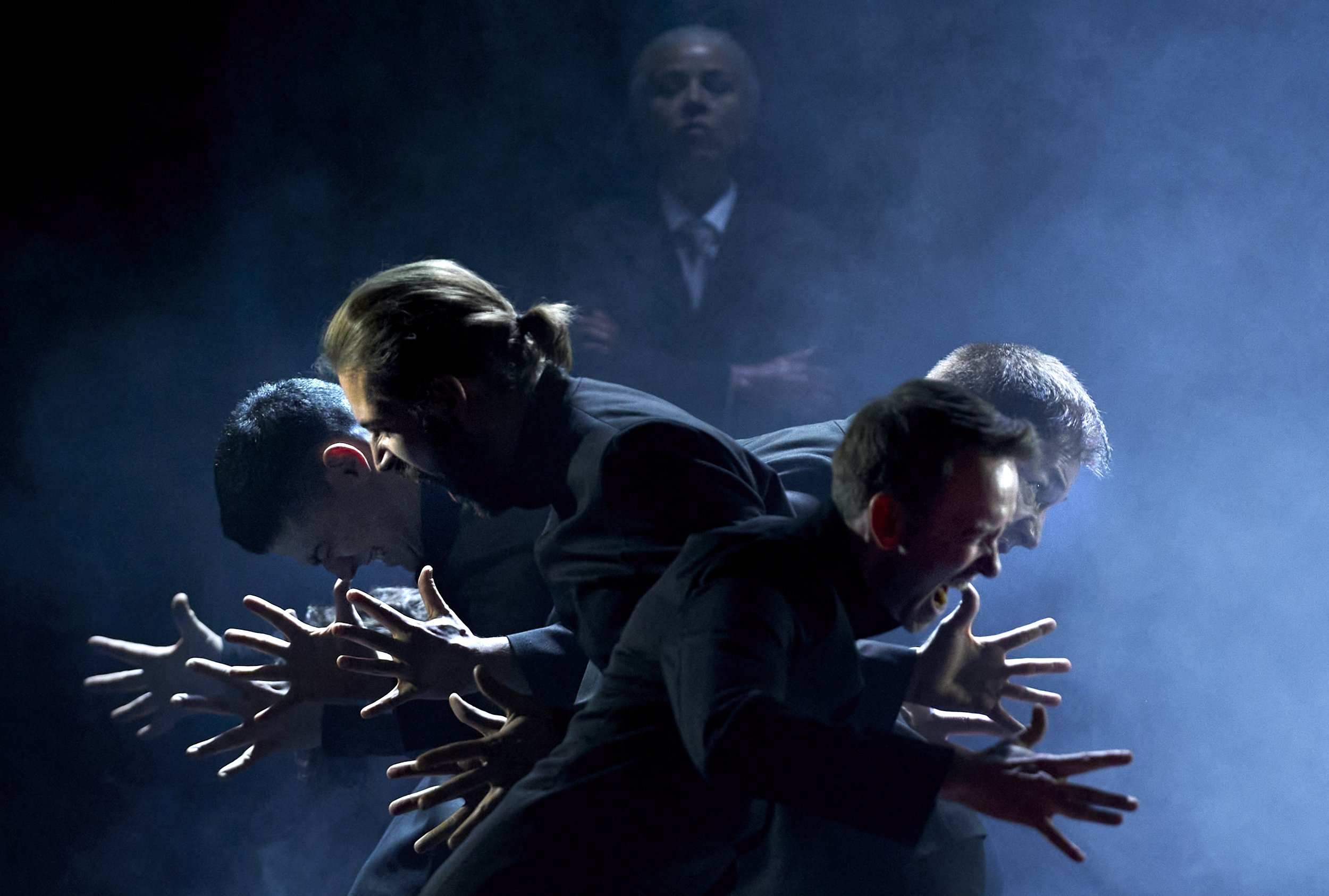 Vier Männer in schwarzen Anzügen mit ausgestreckten Händen, die sich gleichzeitig nach vorne beugen, in einer dunklen, nebligen Umgebung mit einer geheimnisvollen, ernsten Frau im Hintergrund.