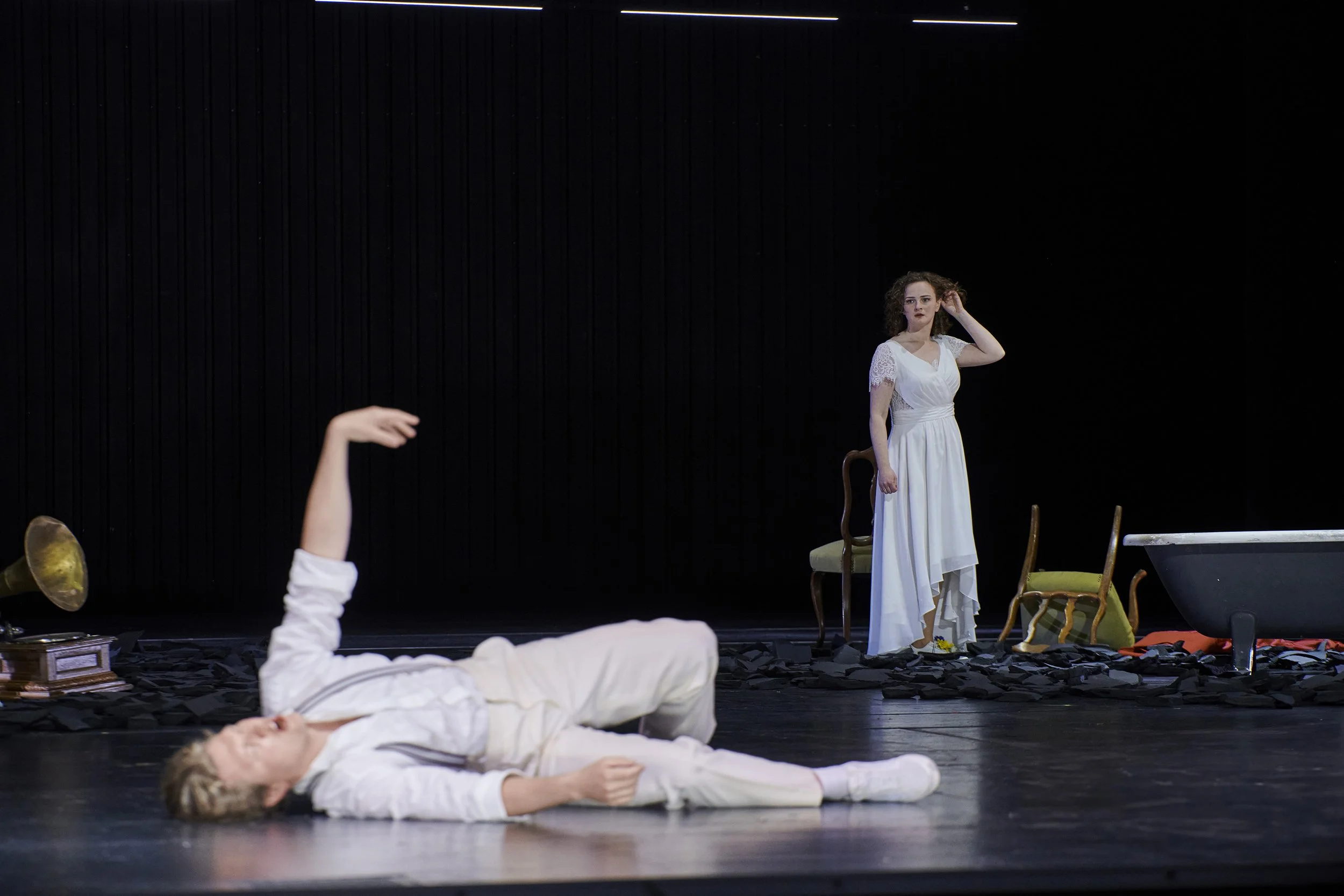 The image shows a stage scene with two women. One is lying down on the floor in white clothing, and the other is standing nearby in a white dress. The scene includes props like chairs, a bathtub, and scattered papers, indicating a theatrical performa