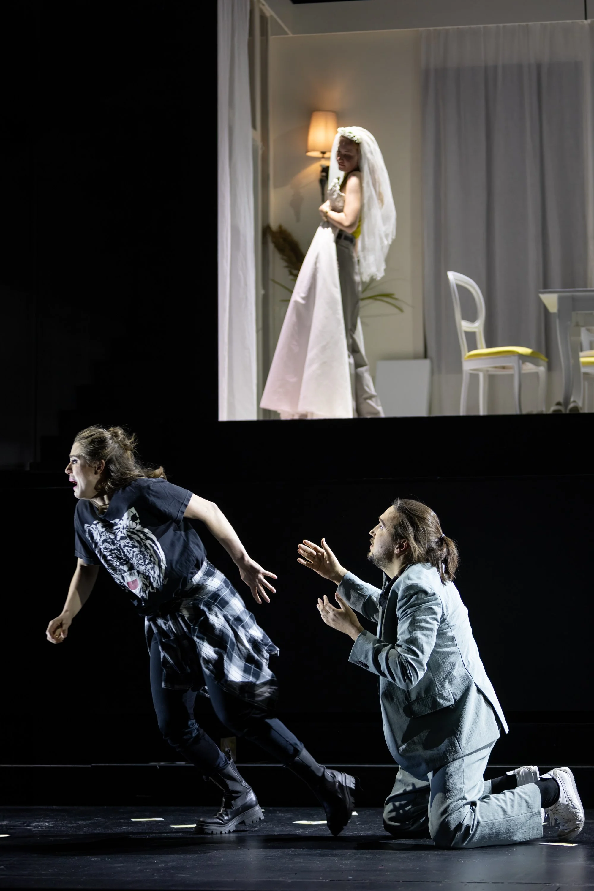 Die Szene zeigt eine Theatervorführung. Im Vordergrund eine junge Frau, die scheinbar in Bewegung ist, während ein Mann auf Knien vor ihr versucht, mit ihr zu kommunizieren. Im Hintergrund auf einer Leinwand ist eine Frau in einem weißen Kleid mit ei