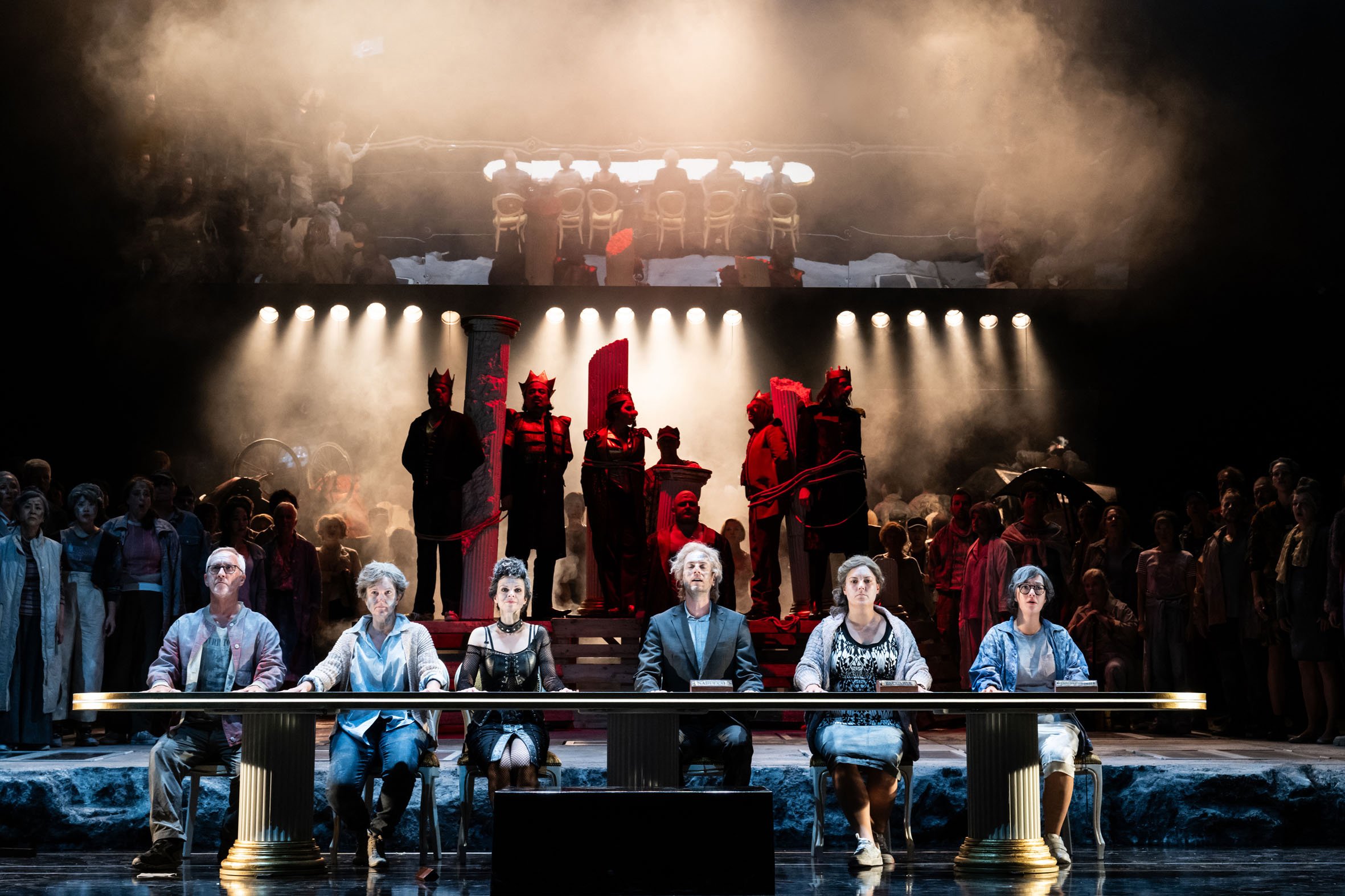 Theateraufführung mit sechs Personen am Tisch im Vordergrund, große Gruppe auf der Bühne im Hintergrund, dunkle Bühne mit dramatischer Beleuchtung, Szenenbild mit antiken Säulen und Figuren, Schauspieler in modernen und historischen Kostümen.