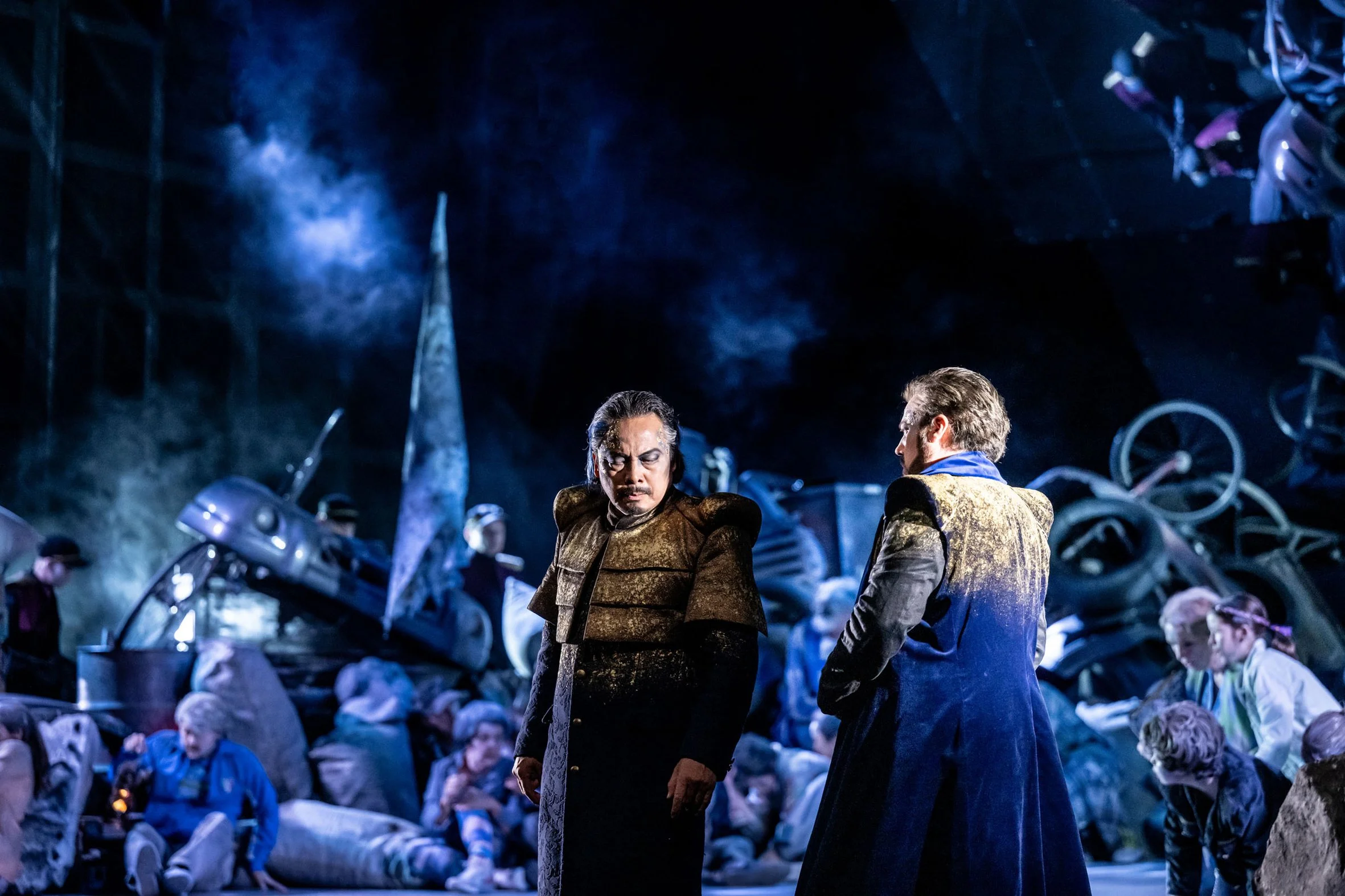 Szene aus einer Oper oder Theaterstück, zwei Figuren in der Mitte in dramischer Haltung, umgeben von weiteren Menschen in einer dunklen, chaotischen Kulisse mit Trümmern und Feuer.