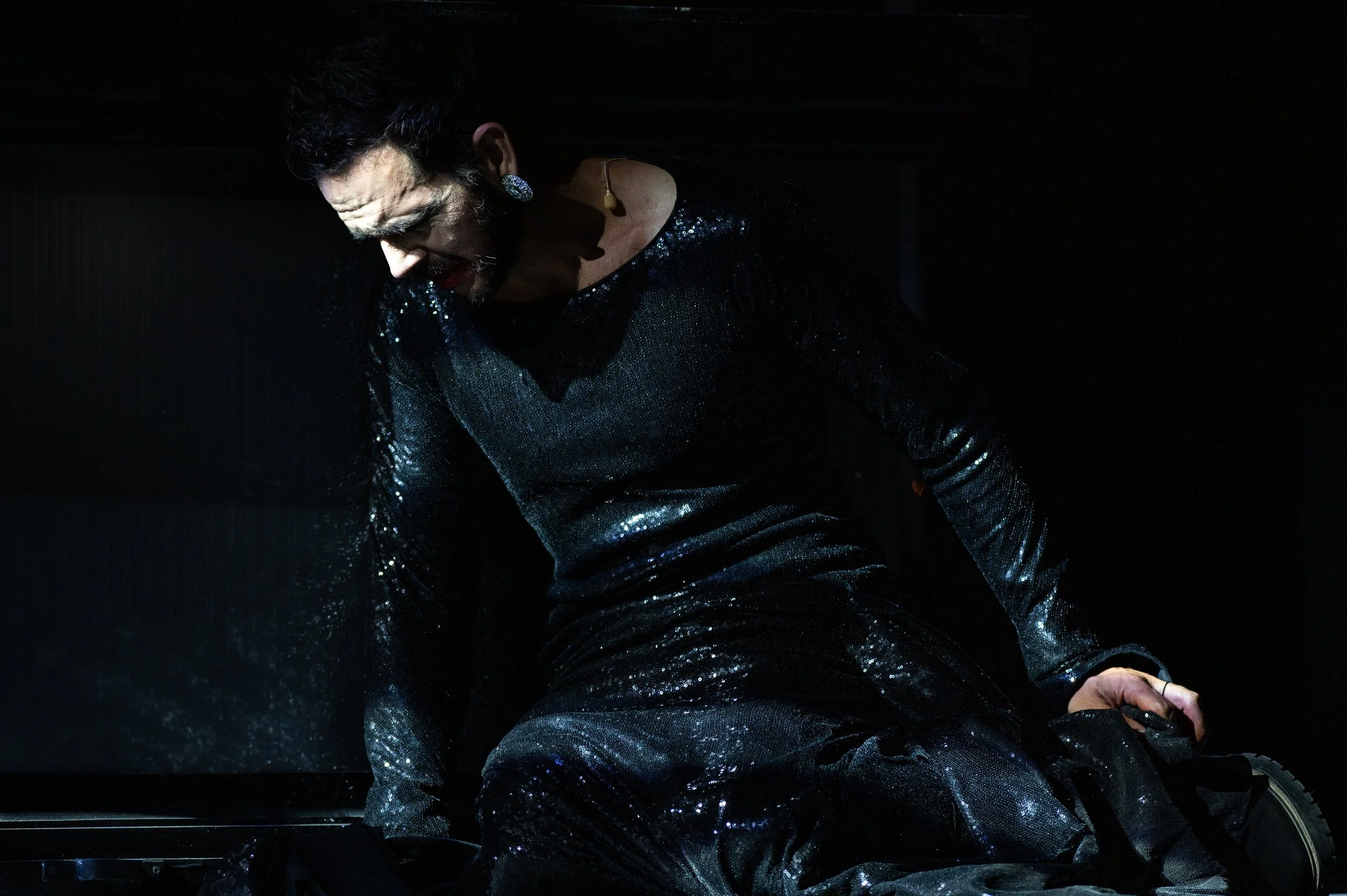 Ein Mann in einem schwarzen, glitzernden Outfit sitzt auf dem Boden und sieht nach unten, während er auf einer dunklen Bühne ist.