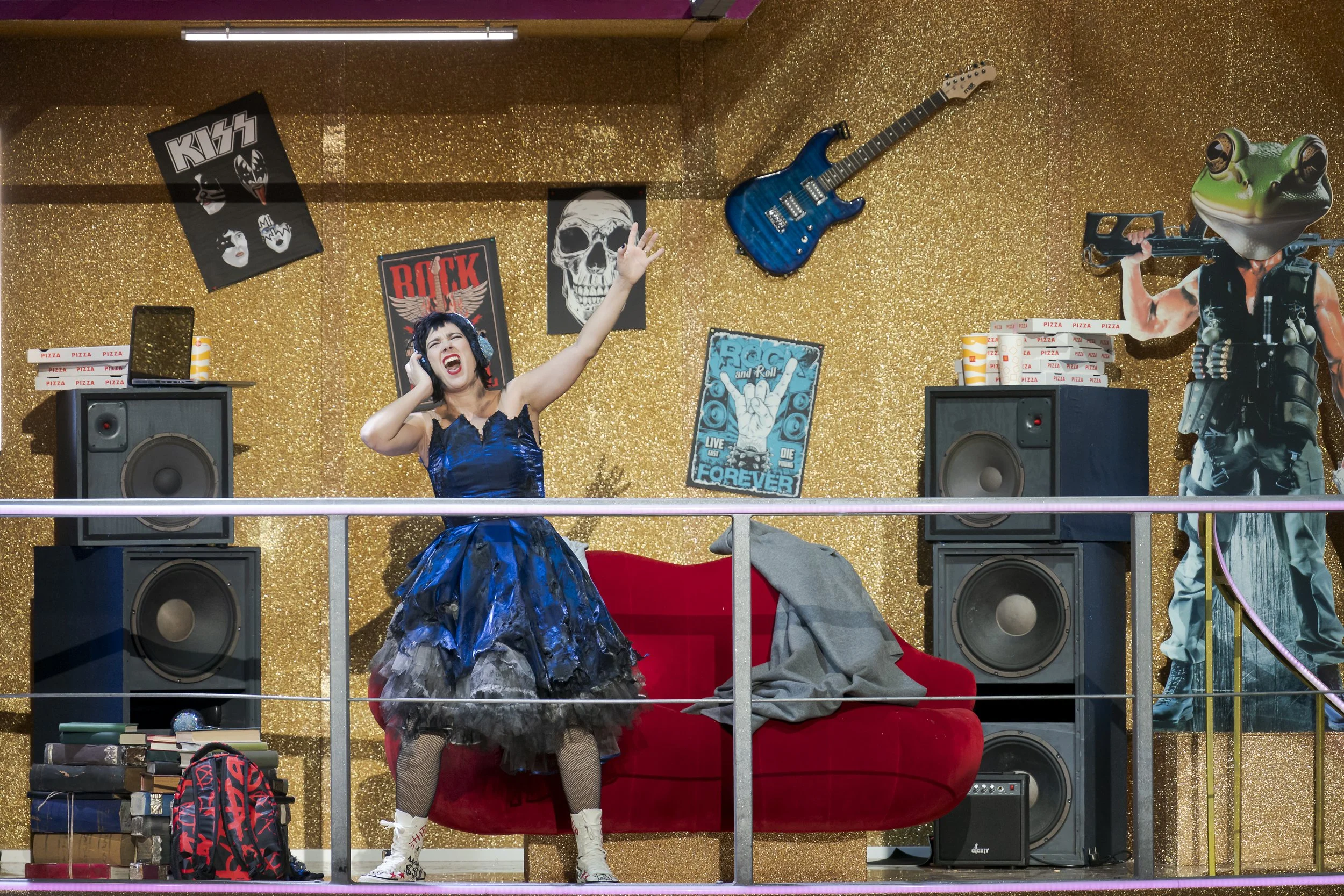 Eine singende Frau trägt ein elegantes, dunkles Kleid und singt mit einem Kopfhörer auf, während sie auf einer Bühne mit goldener deko steht, umgeben von Lautsprechern, Poster mit Rock- und Metal-Motiven, einer Gitarre an der Wand und einem großen Fr