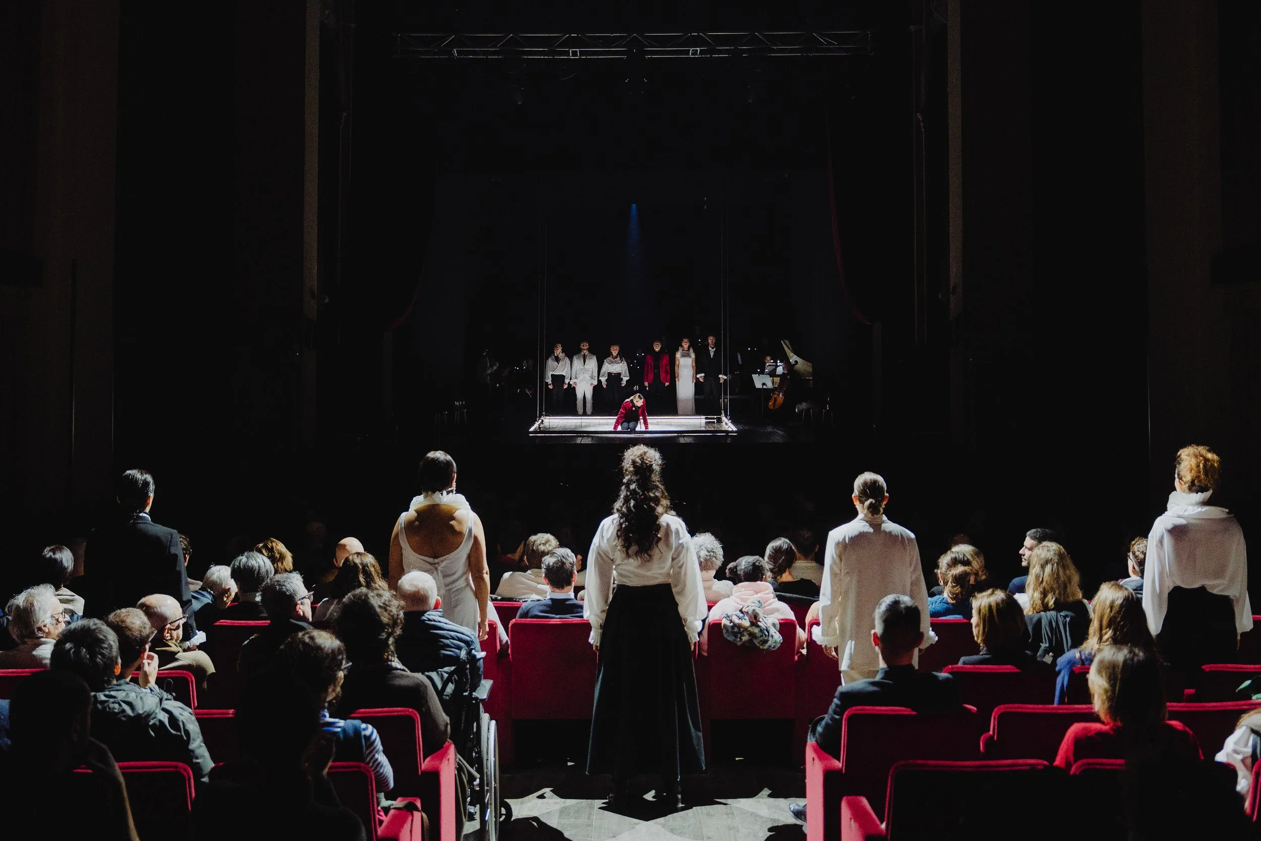 Eine Theateraufführung mit Schauspielern auf der Bühne, im Vordergrund das Publikum in rot gepolsterten Stühlen, einige sitzen, andere stehen, Blick auf die Bühne mit mehreren Darstellern, teilweise in weißen und roten Kostümen, und einem Musiker im 